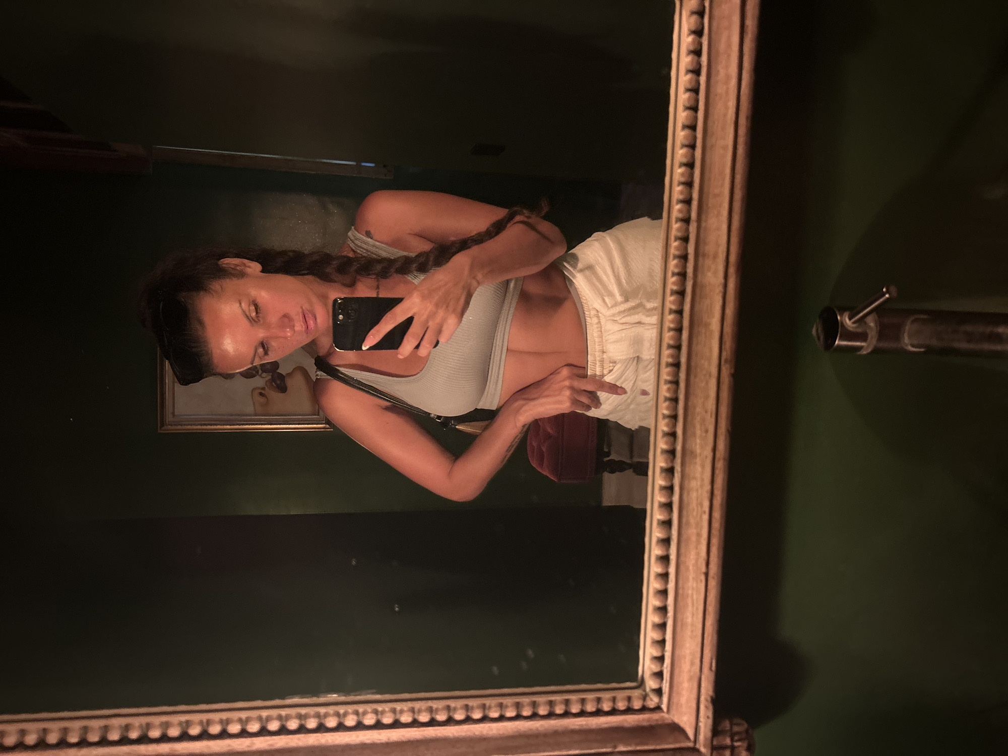 The image is a mirror selfie taken by a woman in North Kuta, Indonesia. She is standing in front of a dark green wall, with a framed mirror in the foreground. The woman is wearing a light blue crop top and white sweatpants. Her hair is pulled back in a braid, and she is holding a smartphone to take the picture. In the reflection, a red velvet stool and a framed painting are visible. The overall lighting is dim, with a spotlight illuminating the woman.