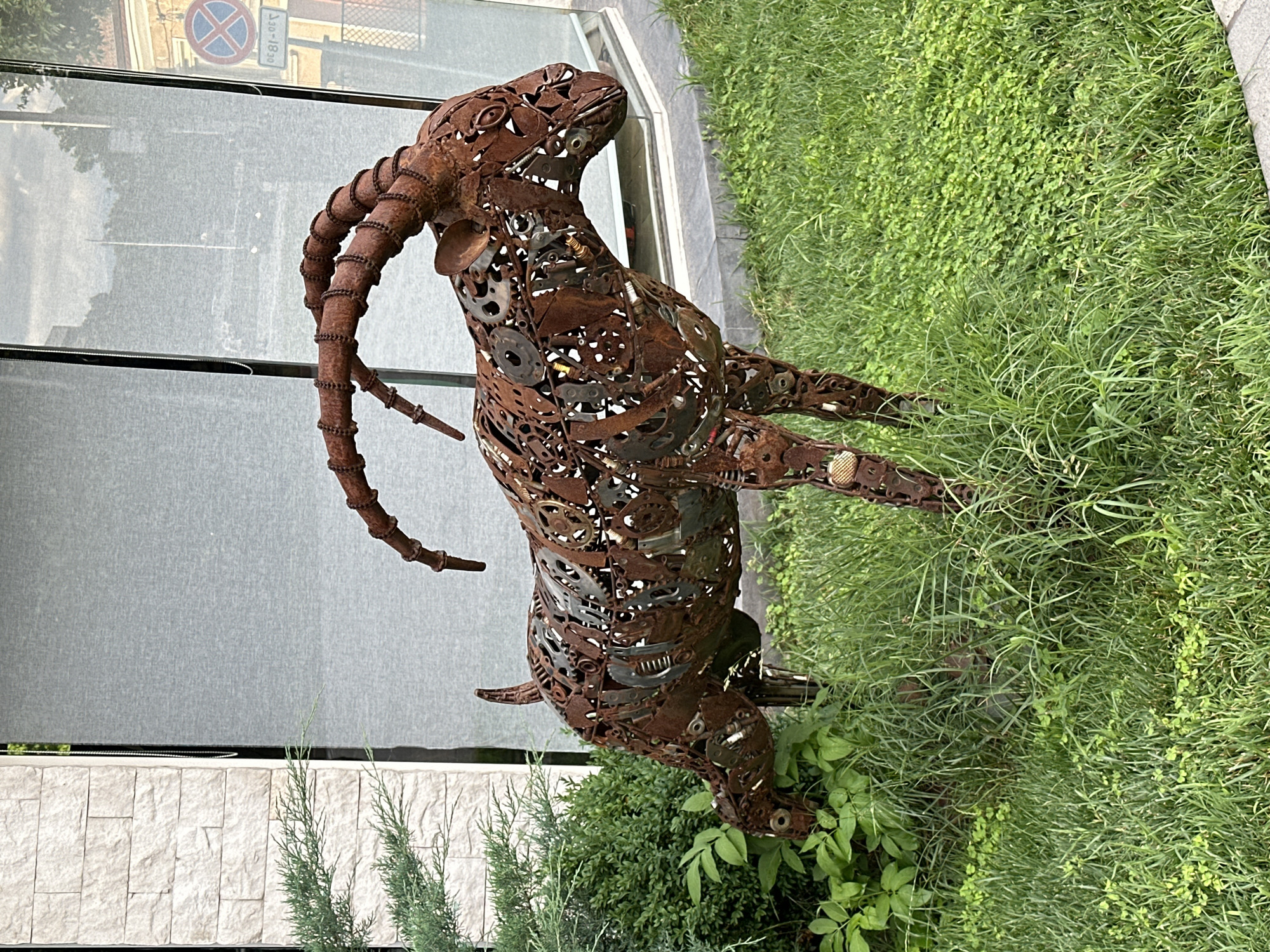 A metal sculpture of a goat, made from recycled metal parts, stands on a patch of grass in front of a modern building with a light-colored brick facade and large glass windows. The sculpture is detailed, with visible gears, bolts, and other metallic components forming its body, horns, and legs. The goat is positioned as if grazing, with its head lowered towards the green vegetation. The background shows reflections in the glass, including what appears to be a street with a circular "no parking" sign. This sculpture is located in the Kapana creative district of Plovdiv, Bulgaria, a pedestrian zone known for its art galleries, craft shops, and vibrant street art.