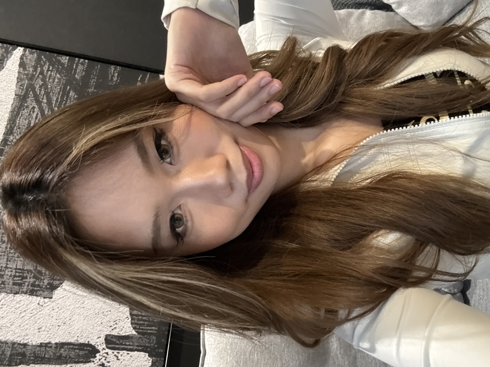 A young woman with long, light brown hair with blonde highlights is reclining with her right hand resting on her forehead. She has fair skin, and green eyes. She is wearing a white jacket over a black top with a zipper. She is indoors. The background includes a black and white patterned surface and a gray and white striped soft surface. There is no evidence in the image to indicate its location as Liangjiang, China.