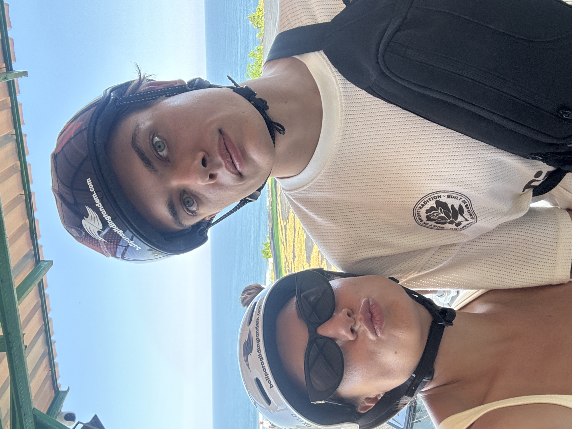 Two individuals, a man and a woman, are taking a selfie outdoors with the ocean and a clear blue sky in the background. The man, positioned slightly behind and to the right of the woman, is wearing a dark helmet with a logo and text that appears to read "baliparaglidingtandem.com". He has striking blue eyes and is wearing a white t-shirt with a circular logo on the left chest. A black bag is slung across his body. The woman, in the foreground, is wearing a white helmet with similar text and logo, sunglasses, and a yellow top. She has her lips puckered for a kiss. The environment suggests a paragliding launch point overlooking the sea. Given the text on their helmets and the scenic ocean view, they are likely in the vicinity of Uluwatu or Padang Padang beach, within Kuta Selatan, Bali, Indonesia, a popular area for paragliding activities.