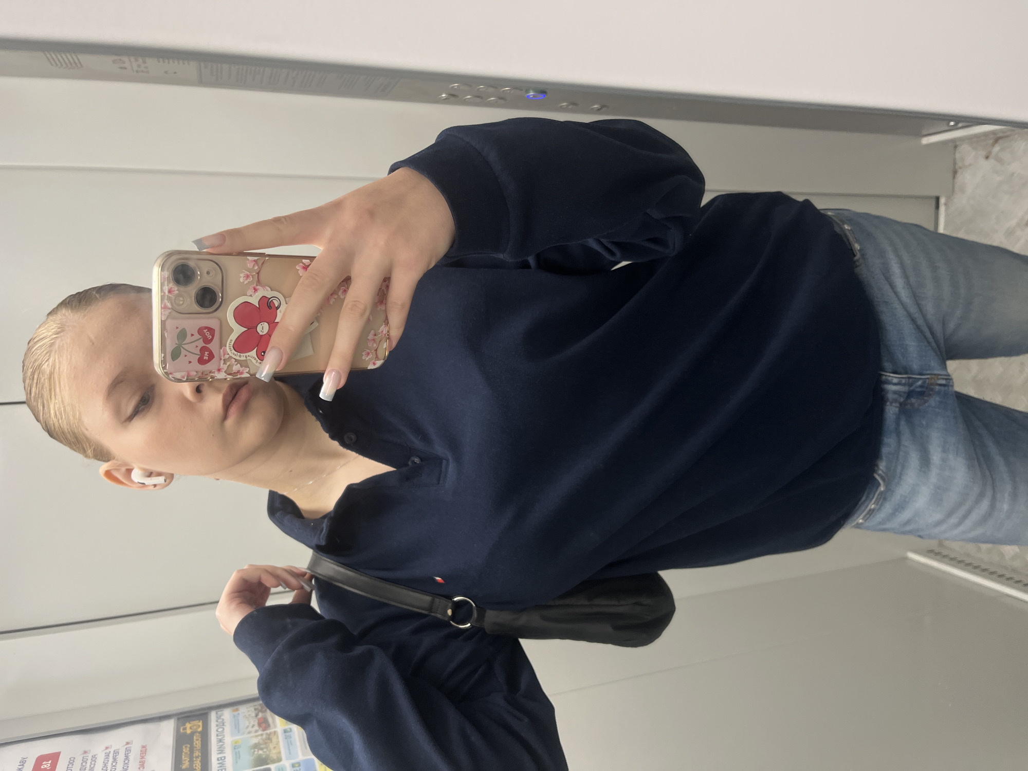 A young woman with blonde hair pulled back in a bun is taking a mirror selfie in what appears to be an elevator. She is wearing a navy blue collared long-sleeved shirt and light wash denim jeans. She has on white wireless earbuds and is holding her smartphone, which has a case decorated with cherries and flowers. She is also wearing a small silver necklace. A black purse with a silver hoop is slung over her left shoulder. The elevator walls are a neutral gray, and a metallic panel with buttons is visible to her right. A sign with text in Russian is partially visible on the wall to her left. The overall lighting suggests an indoor setting. There is no indication of the location being in the city of Perm, Russia.