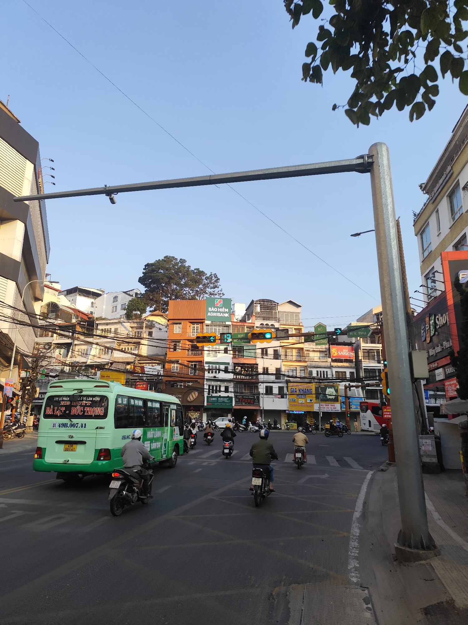 This image shows a street scene in Da Lat, Vietnam. The intersection is busy with traffic, including a green bus, several motorcycles, and pedestrians. Numerous buildings with varying architectural styles line the street, including some with visible signs for businesses like Agribank, Gia Khang Mobile, and Cafe. Overhead, traffic lights display green signals, and a metal traffic signal structure spans the width of the road. The sky is clear and blue.