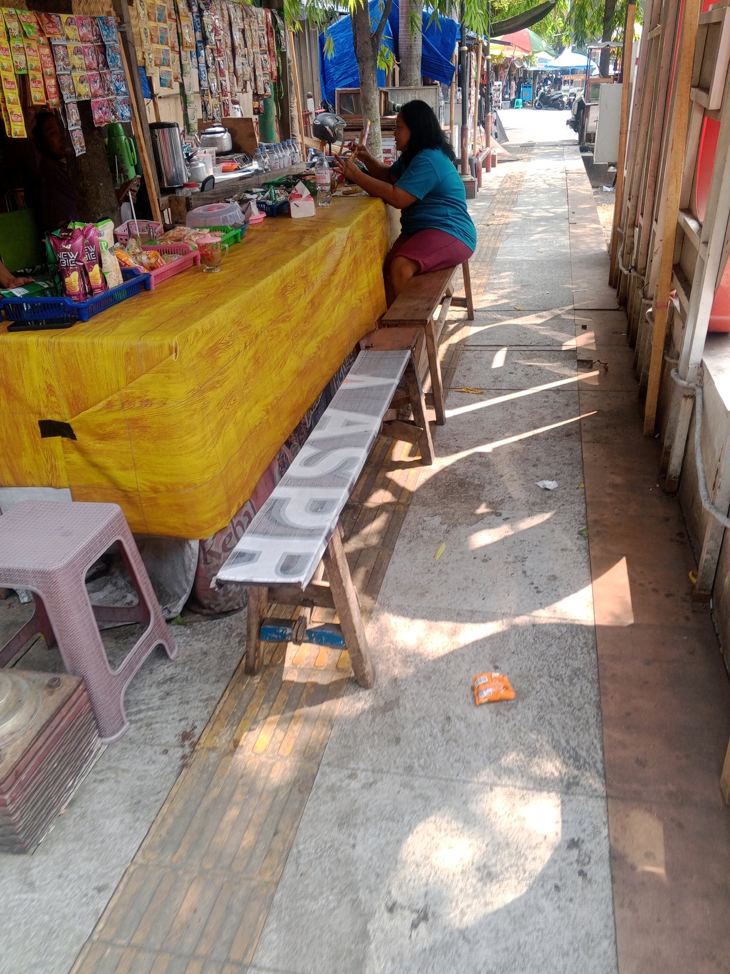 A woman wearing a turquoise t-shirt and maroon shorts sits on a wooden bench in front of a street-side food stall. She is holding a mobile phone in her left hand and looking at it. The stall is covered with a yellow tablecloth, displaying various snacks and drinks. In the background, there is a street lined with stalls and trees, with a blue tarp and some tents visible. The ground is paved with concrete, and sunlight casts shadows across the scene. The image appears to be taken in a public area in Wonogiri, Indonesia.