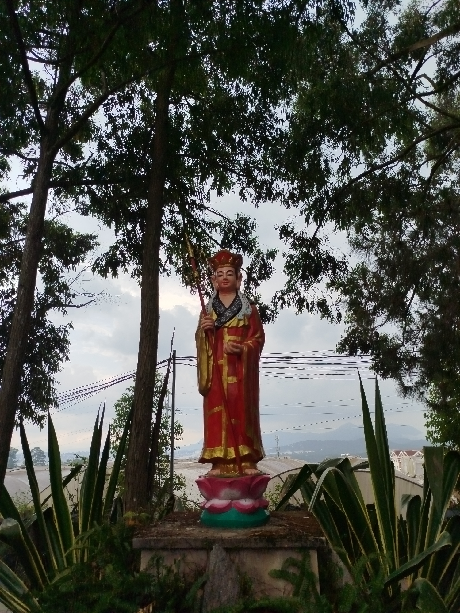 A statue of a robed figure with a staff stands on a lotus base in a garden setting. The figure is adorned in red and gold garments with a distinct headdress. Trees frame the statue, and in the background, there are views of Xuân Trường - Đà Lạt, Vietnam including buildings, mountains, and an overcast sky. Agave plants flank the statue's base. Powerlines can be seen behind the statue and above the treeline.