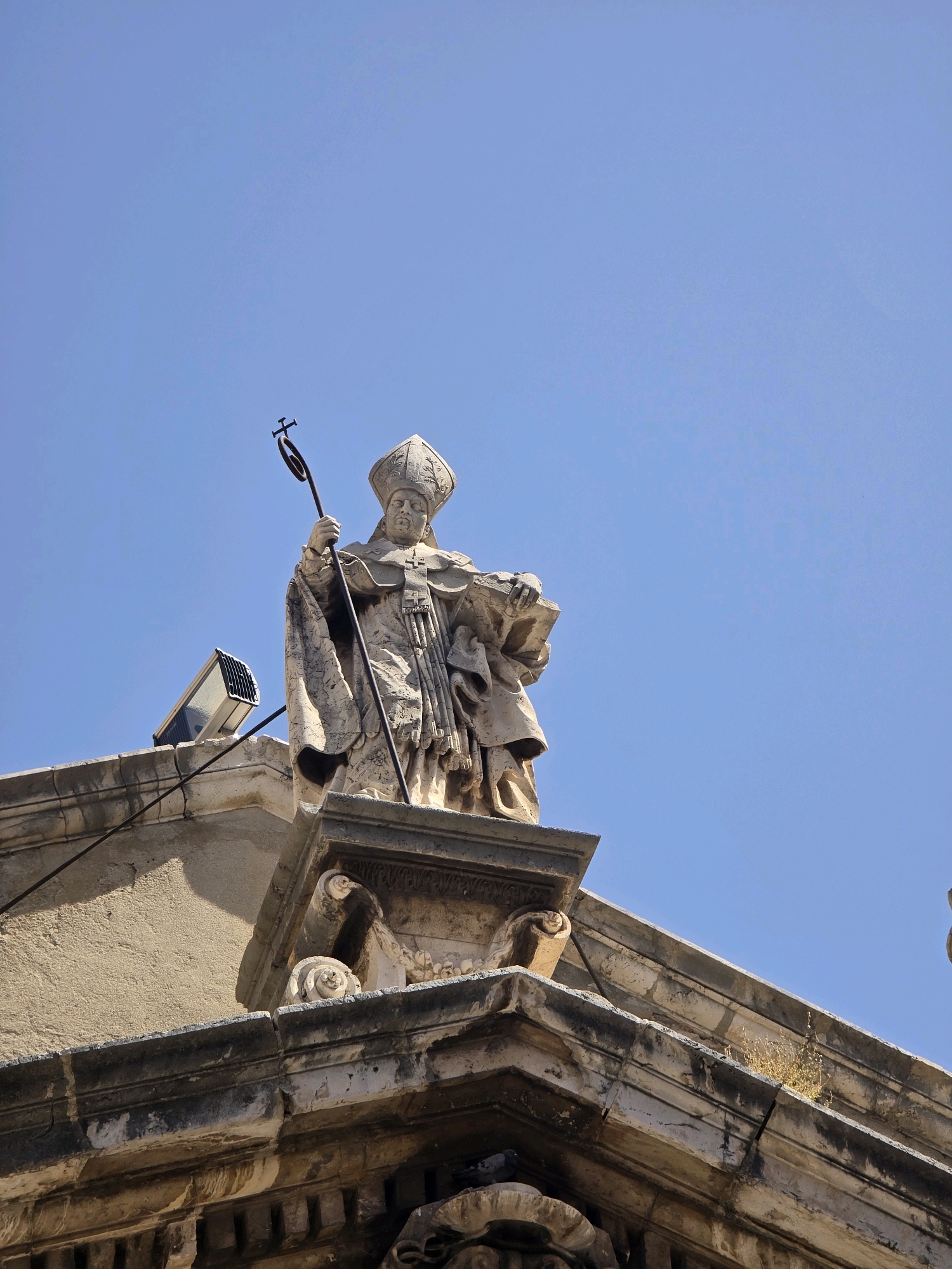 A stone statue of a robed bishop, wearing a miter and holding a crozier and a book, stands atop a building against a clear blue sky. The statue is weathered, and the building appears to be an old stone structure with ornate architectural details. A modern floodlight is mounted on the building to the left of the statue. The image was taken in Jaén, Spain.