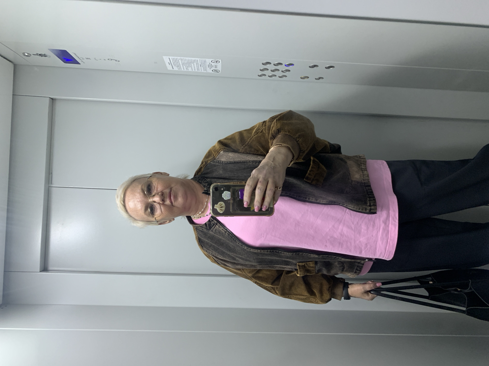 An older woman is taking a selfie in an elevator. She has white hair, glasses, and is wearing a pink shirt with a brown jacket, along with black pants. She's holding a phone with stickers on the back in her right hand, and a black bag in her left hand. The elevator has a gray interior with buttons and indicators on the wall. The location is likely Chaikovsky, Russia.