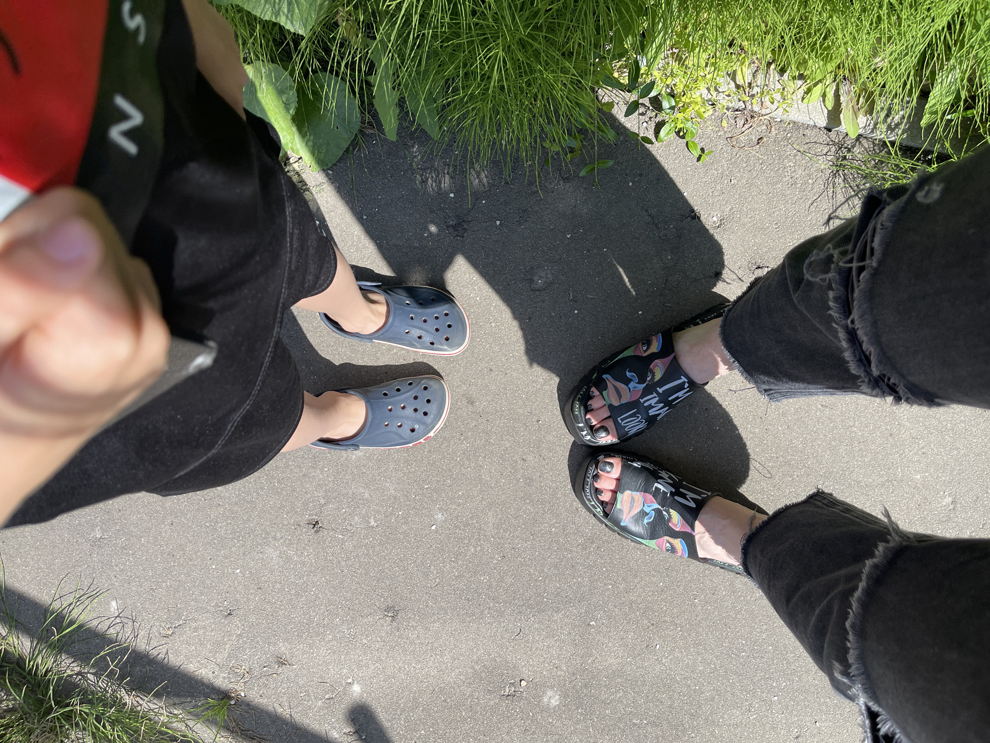 An overhead, eye-level shot shows two pairs of feet and legs standing on a concrete path next to a grassy area. The person on the right is wearing blue Crocs and dark shorts, and their leg is bent. The person on the left is wearing black slides with a colorful face print and ripped black jeans. They are both standing with their feet facing forward. The scene is illuminated by sunlight, creating shadows on the path. The location is the city of Kommunarka, Russia.