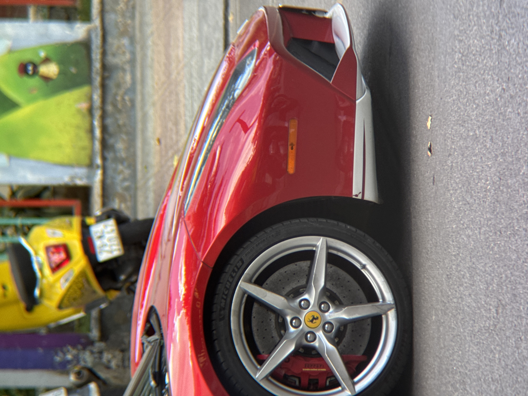 The image is a close-up, low-angle shot of the front passenger side of a red Ferrari, parked on an asphalt street in Nha Trang, Vietnam. The car's distinctive silver five-spoke wheel, with the Ferrari logo at its center and red brake calipers visible behind it, is prominently featured. The lower portion of the car's aerodynamic bodywork, including its front bumper and a small orange indicator light, is also visible. In the background, slightly out of focus, is a bright yellow scooter with a license plate, and a colorful mural depicting abstract green shapes with a small figure. The overall scene suggests an urban environment, with the red Ferrari being the primary subject.