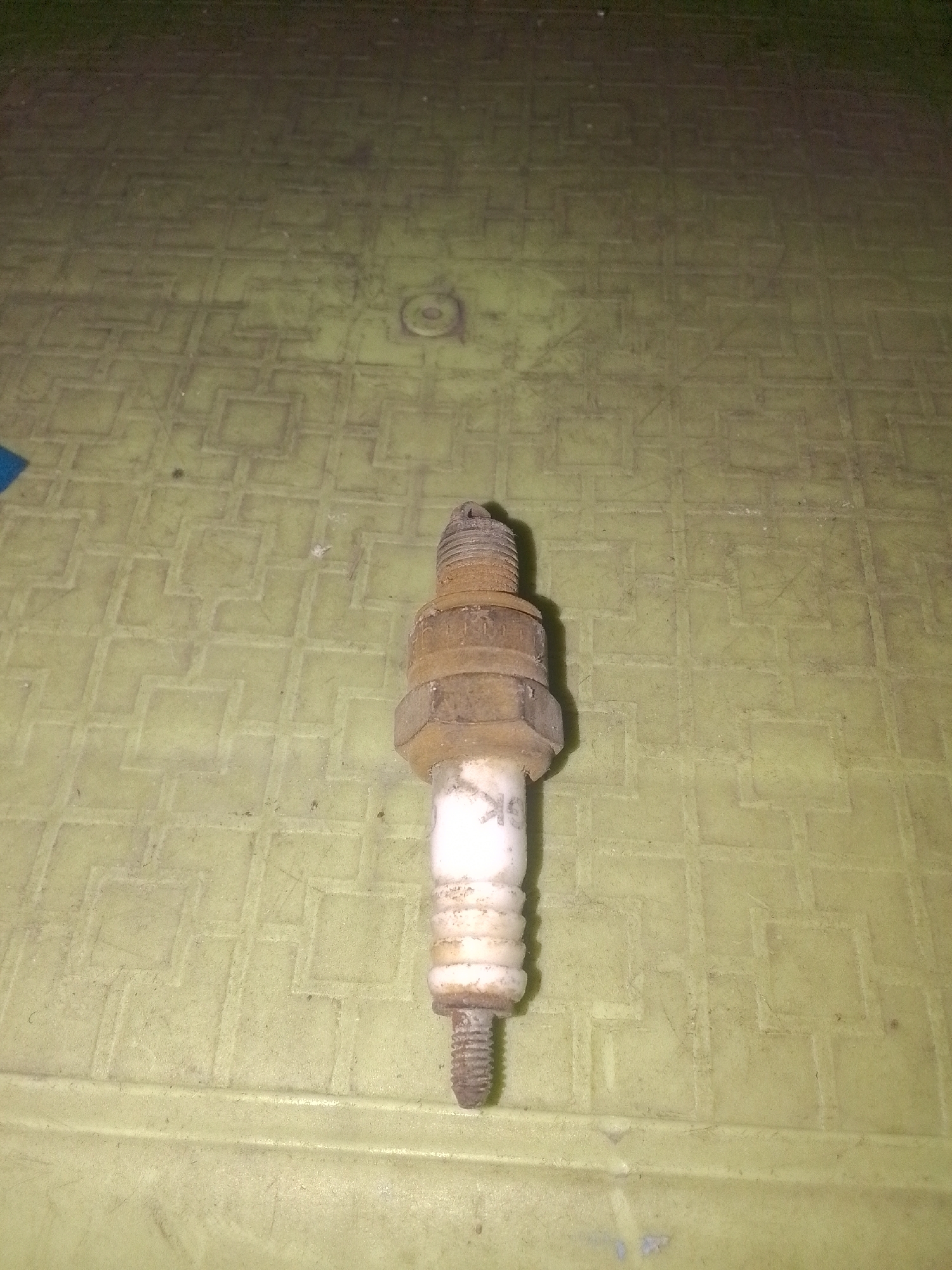 A single spark plug is centrally placed on a textured, light-green surface. The spark plug appears used and is slightly dirty. The background is a uniform, slightly blurred light green. The overall lighting is dim. There is no indication of the city of Gunungmasigit, Indonesia in the image.