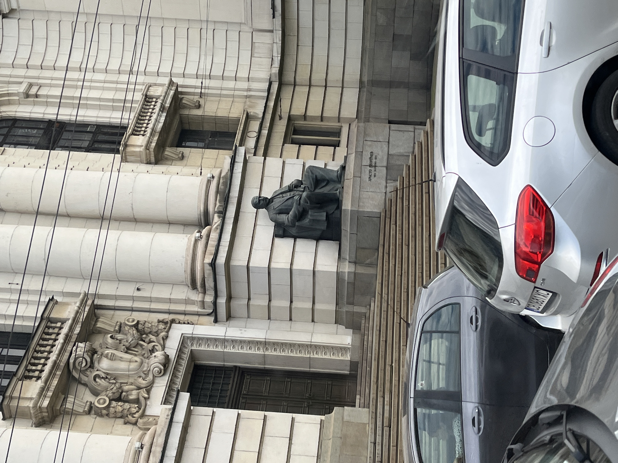 The image shows a sculpture of a seated man on the exterior of a grand, classical-style building. The sculpture appears to be made of dark stone and is positioned on a tiered platform. Above and to the left of the sculpture, there are windows with dark panes set into the stone facade. Decorative architectural elements, including ornate carvings and columns, are visible on the building. The building's exterior is made of light-colored stone blocks and features intricate stonework.
In the foreground and to the right of the building, several cars are parked. One car is silver and has its rear visible, showing a red taillight and a license plate with text that is partially legible. Another car is dark gray and is parked behind the silver car. A third car, also gray, is partially visible in the lower right corner. The cars are parked on a paved surface adjacent to the building.
The scene appears to be outdoors, and the lighting suggests it is daytime, though the weather is not clearly indicated. There is no discernible activity taking place other than the static presence of the sculpture, building, and parked cars.
Visible text on the sculpture base reads: "ХРИСТО БОТЕВ" and some numbers which are difficult to read. The license plate on the silver car has text that appears to be "CB 0661 KC".