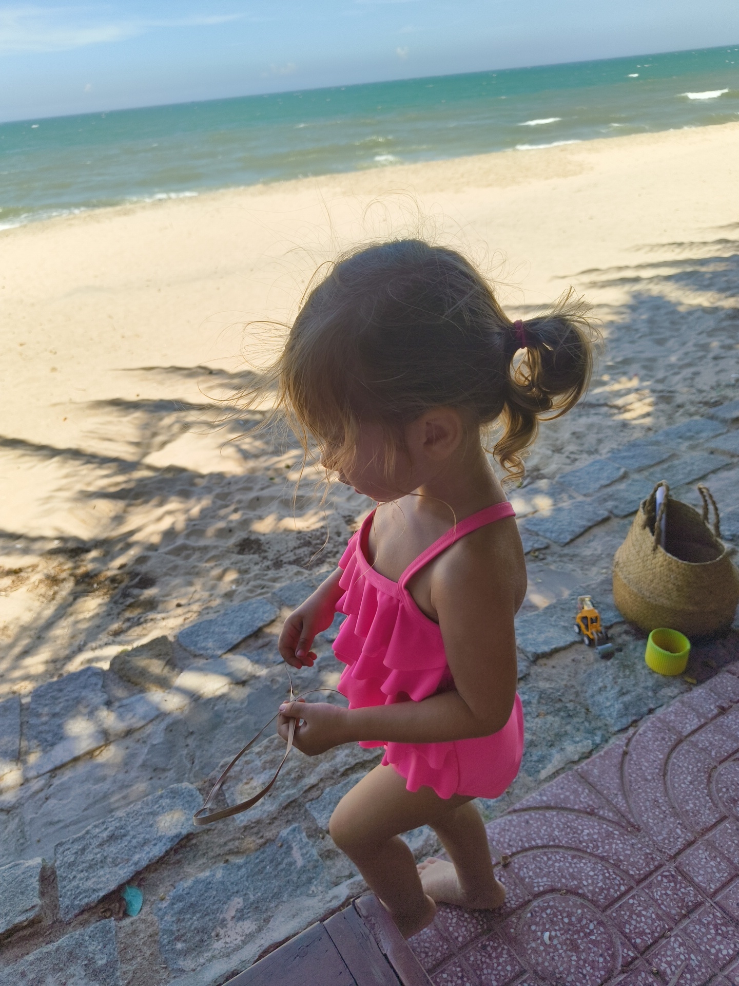 In Phu Thuy Ward, Vietnam, a young girl with blonde hair tied in a ponytail wears a bright pink ruffled swimsuit. She is standing on a stone pathway next to a sandy beach. The girl is holding what appears to be a pair of sunglasses or a thin object in her hands. To her right, a woven straw bag sits on the pathway, next to a small yellow toy truck and a bright green cup. The background features a sandy beach transitioning into the ocean under a clear blue sky with some clouds. The sun casts dappled shadows from palm fronds onto the sand and pathway.