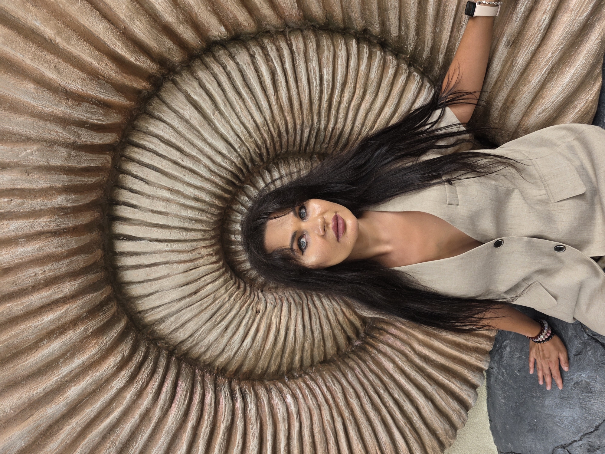 A woman with long, dark hair and blue eyes is seated in front of a large, textured, shell-like structure. She is wearing a beige vest with two dark buttons. Her left hand rests on a dark, rock-like surface, adorned with bracelets. Her right arm is extended, and she is wearing a smartwatch on her wrist. The background structure is predominantly brown and beige, with a ribbed, spiral pattern. The overall setting appears to be indoors, possibly a museum or an art installation, and the environment is well-lit. The location is identified as Estosadok, Russia.