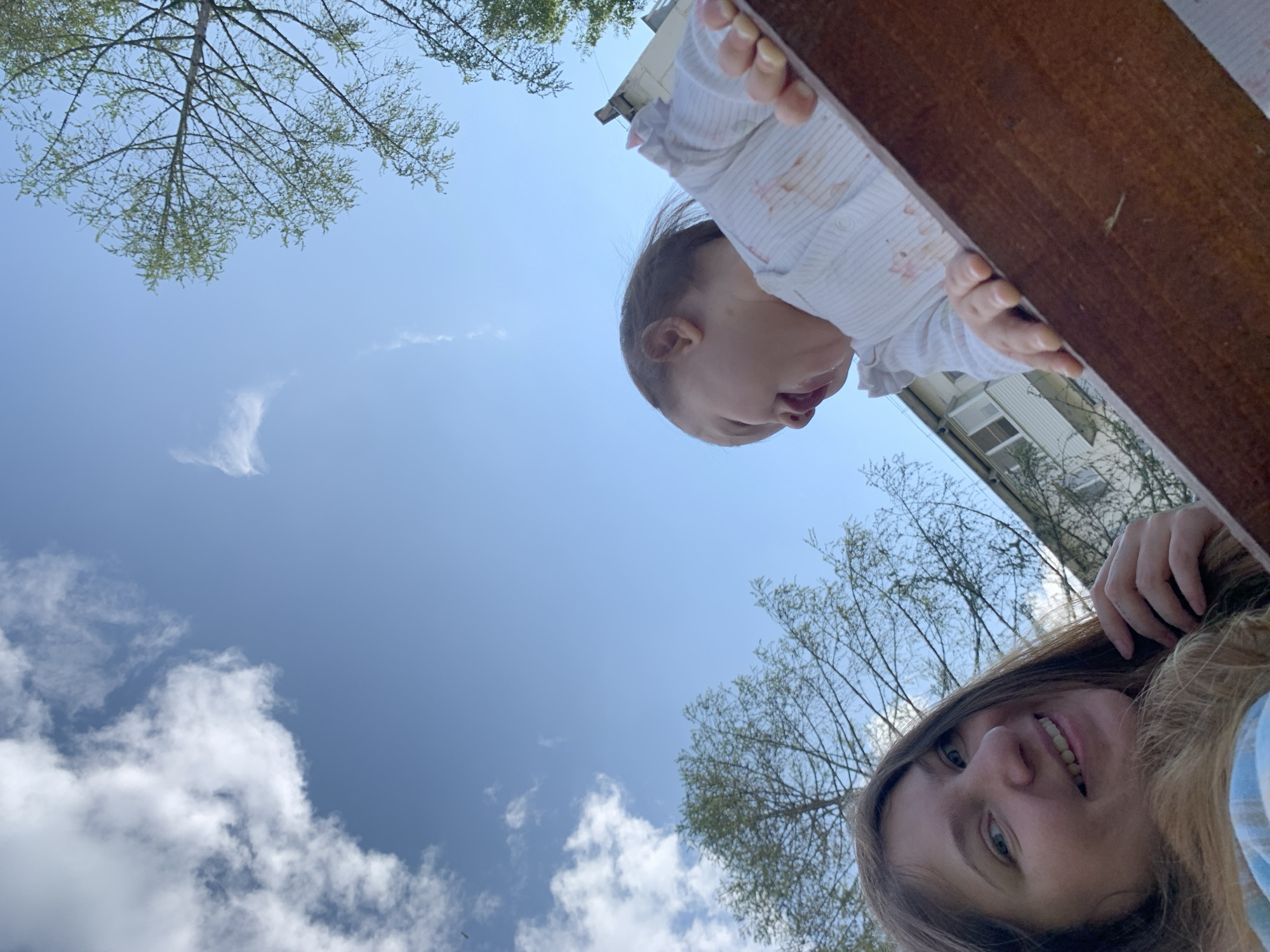 An outdoor, low-angle, medium shot features a woman and a toddler against a backdrop of blue sky with scattered clouds, trees, and a multi-story building. The woman, positioned on the left, has long, brown hair, fair skin, and is smiling. She is wearing a blue and white plaid shirt. The toddler, on the right, is wearing a white striped shirt with pink accents and is looking to the left with their mouth open. Both are leaning over a brown wooden structure. The building appears to be residential, with windows and balconies visible. The overall scene suggests a casual moment outdoors in the city of Chaikovsky, Russia, possibly on a playground or balcony.