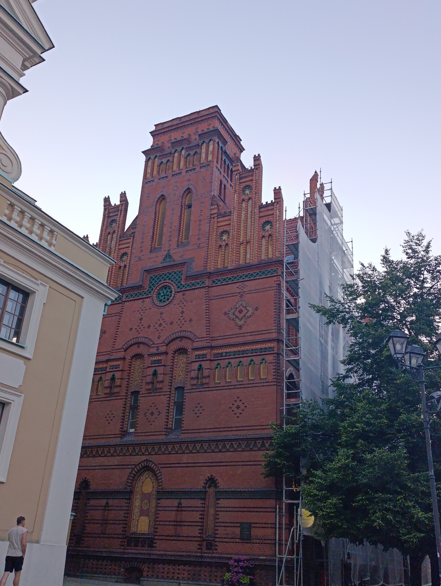 The image shows the Neo-Gothic architecture of the Toruń Town Hall, located in Toruń, Poland. The building is constructed of red brick and features intricate decorative elements, including arched windows and a central clock. A portion of the building on the right is covered in scaffolding and white sheeting, suggesting ongoing renovation or construction work. A large green tree is positioned to the right of the building, partially obscuring the scaffolding. Two streetlights with multiple bulbs are visible near the tree. In the foreground on the left, a portion of a lighter-colored building with classical architectural elements is visible. A man wearing a light-colored t-shirt and dark shorts stands facing away from the camera on the left side of the frame, near the lighter building. The sky above is clear and pale, indicating it is likely early morning or late evening.