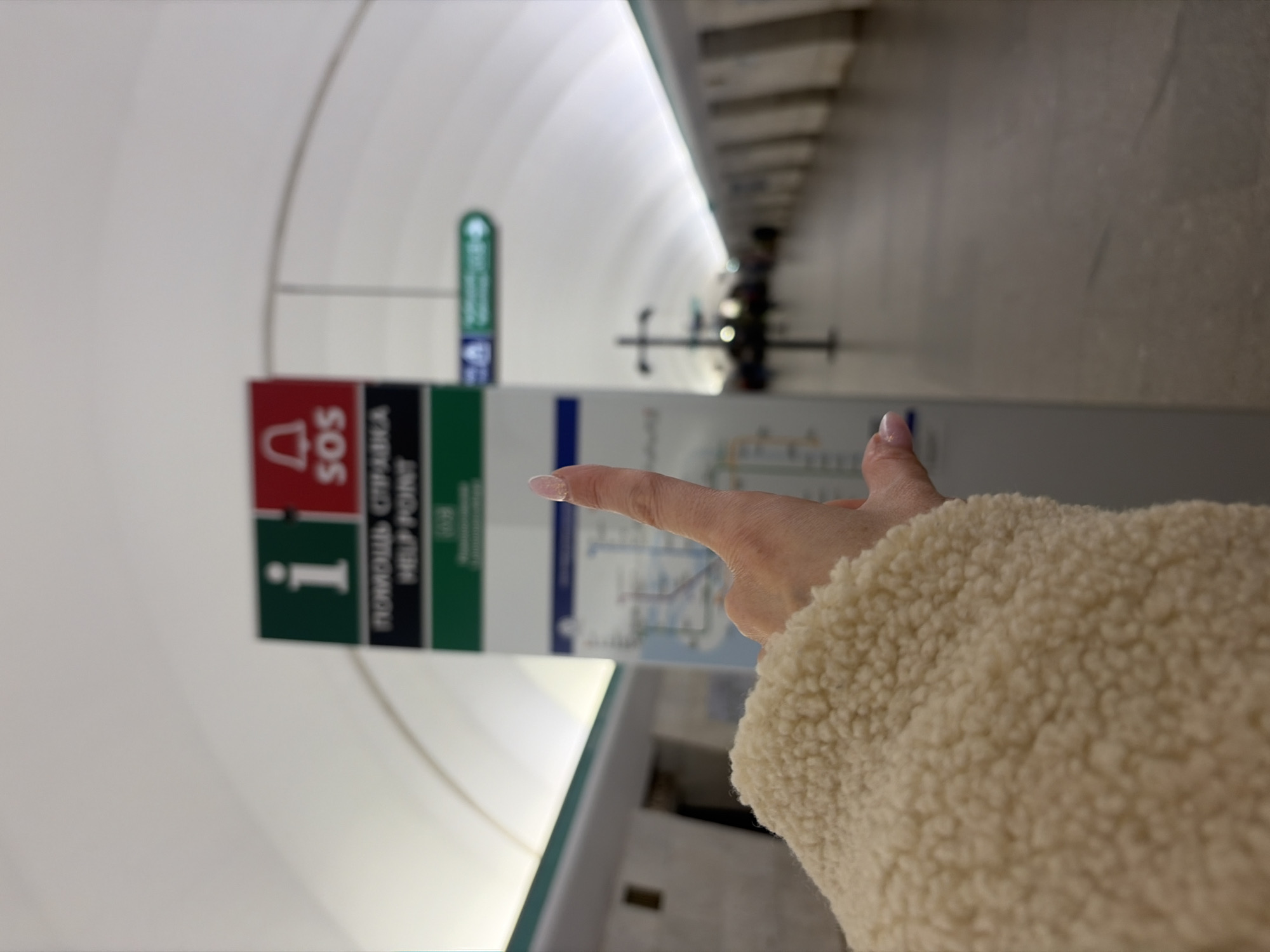 The image shows the platform of a Saint Petersburg metro station, likely one of the newer stations with a modern design, given the curved white ceiling and the information pillars. The pillars contain a red "SOS" sign, a green "i" information symbol, and a metro map. The text "ПОМОЩЬ СПРАВКА" (help reference) and "HELP POINT" are visible on one of the pillars. A person's hand is pointing towards the metro map. The person is wearing a cream-colored fleece jacket. In the background, a green station sign is visible, along with benches for passengers to sit on.
