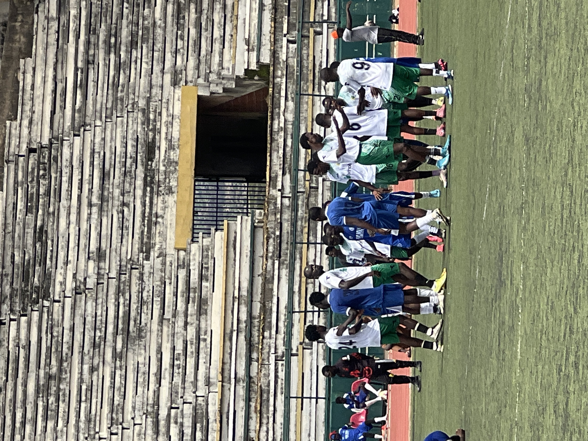 In the New Stadium, Nigeria, a group of young men in green and white soccer uniforms huddle together on a green turf field, their faces obscured or turned away. Their athletic stance suggests a moment of strategy or camaraderie. In the background, weathered concrete bleachers rise up, stained with age and neglect, contrasting with the vibrant green of the field. A yellow-painted doorway with a metal grate hints at the stadium's infrastructure. A few other individuals in athletic wear and casual attire are scattered around the stadium periphery, some seated on red plastic chairs, others standing. The overall atmosphere is one of organized athletic activity within a somewhat dilapidated but functional sporting venue.