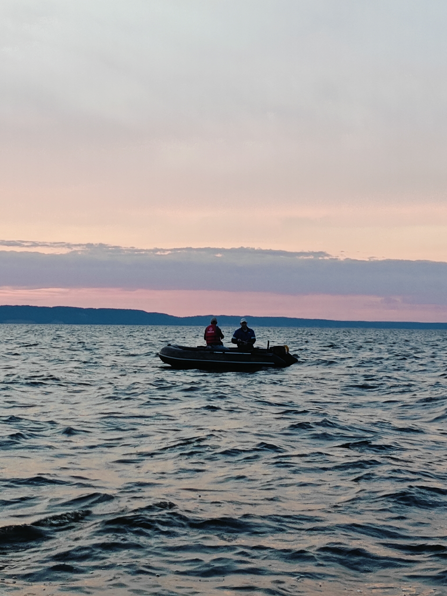 The image depicts two individuals in a black inflatable boat on a body of water. The sky above is a gradient of pale pink and gray, with horizontal bands of clouds. A distant, dark landmass forms the horizon line. The water is choppy, with visible ripples and waves. The person on the left is wearing a red vest and a dark cap, while the person on the right wears a white cap and a blue shirt. The boat appears to be stationary. The scene suggests a calm, possibly dusk or dawn setting. The location is identified as Stepanovo, Russia.