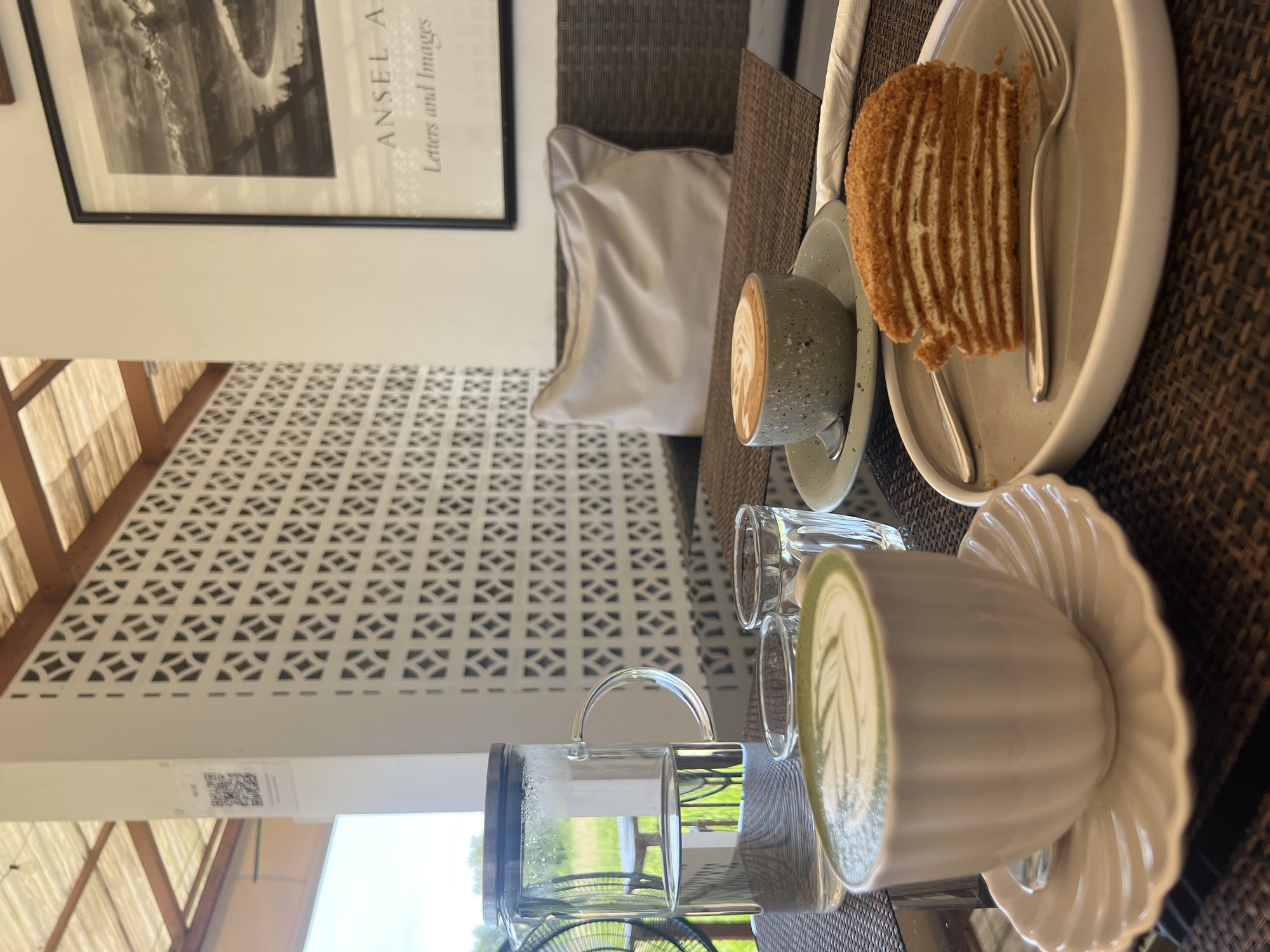 This image depicts a serene outdoor cafe setting in Baan Mae Nam, Thailand. In the foreground, a white ribbed teacup filled with a matcha latte, complete with leaf art, rests on a matching saucer. To its right, a slice of honey cake, characterized by its thin layers and golden-brown crust, is presented on a pale grey plate with a fork. Behind these treats, a small glass of water and a larger glass carafe filled with water are visible. In the mid-ground, a grey speckled mug containing a cappuccino with latte art sits on a saucer. A woven grey cushion is placed on a dark rattan sofa in the background. The cafe's ambiance is enhanced by a distinctive white patterned wall and a wooden slatted roof. The scene is illuminated by natural light, suggesting it's daytime.