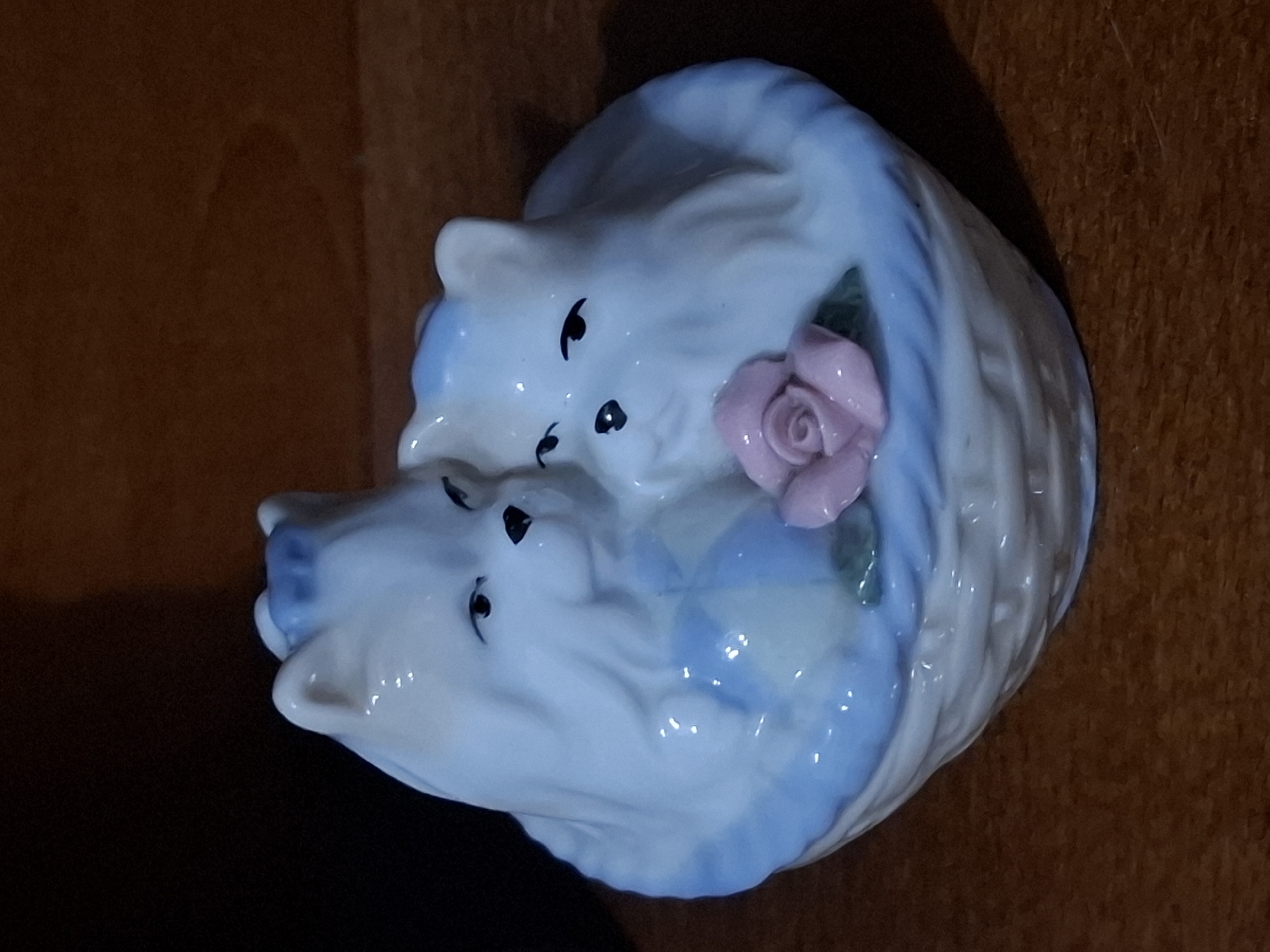 A ceramic figurine of two white kittens nestled in a blue-trimmed, woven basket is displayed against a dark, blurred background. A pink rose with green leaves sits in the foreground of the basket. The figurine is illuminated from above and to the left, casting a shadow to the right. The surface beneath the figurine appears to be a polished wooden table. The location in Almaty, Kazakhstan, is not discernible from the image.