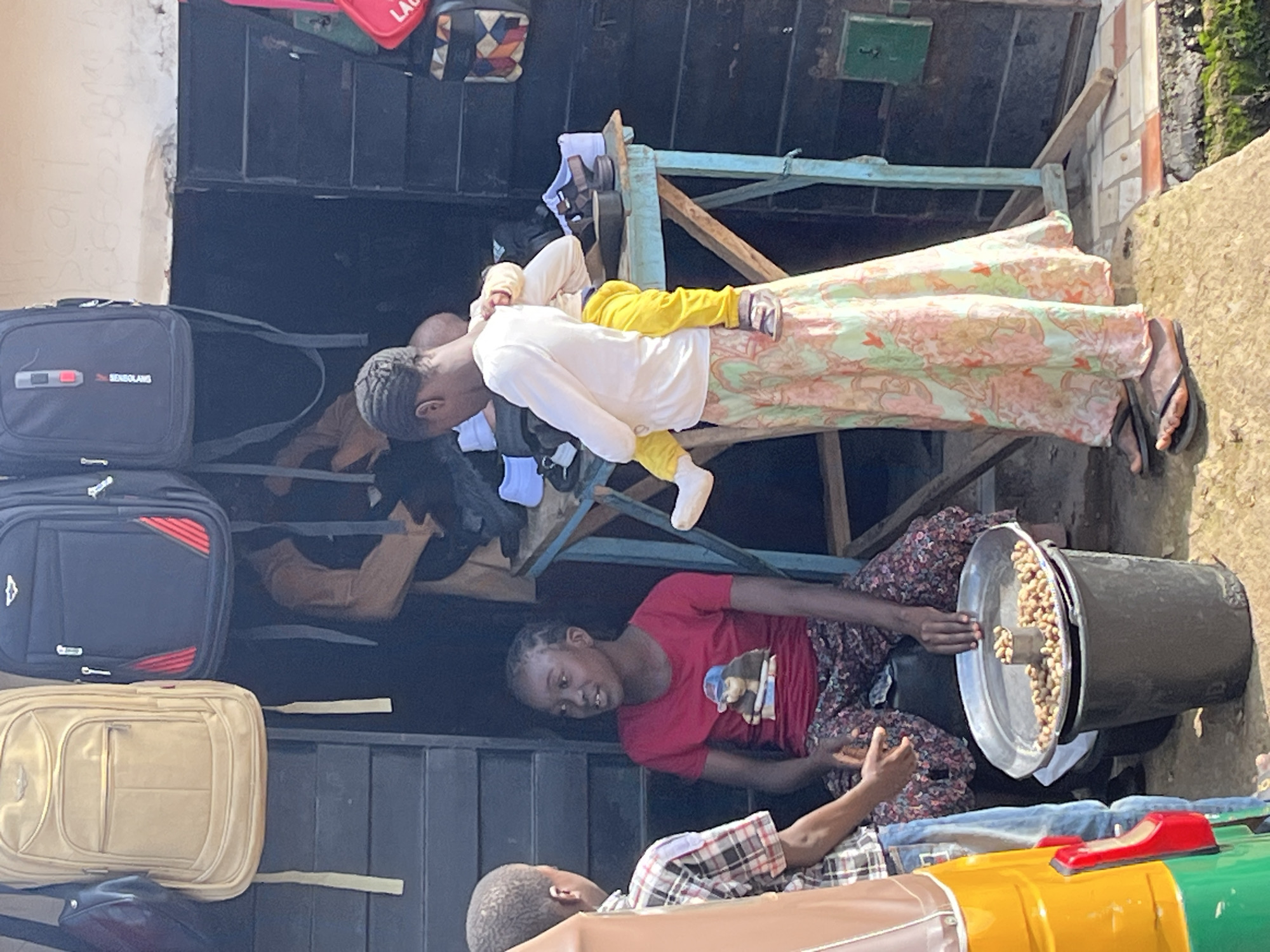 The image depicts a street scene in Jos, Nigeria, focusing on a small market stall. In the foreground, to the left, a person in a plaid shirt and jeans stands next to the side of a brightly colored auto-rickshaw. To the right of the auto-rickshaw, a young girl wearing a red t-shirt with a graphic print and patterned pants sits. In front of her is a large black bucket topped with a metal tray filled with peanuts. A woman stands behind the girl, carrying a baby on her back. The baby is wearing yellow pants and a white shirt. The woman is wearing a long, patterned skirt and a white long-sleeved shirt. Behind them, the market stall is visible, with backpacks hanging from the top and shelves laden with various items, including shoes. The background shows a wooden structure with shelves and some signage. The ground is paved with tiles in some areas and dirt in others, with some greenery visible. The overall impression is a bustling, everyday scene at a local market.