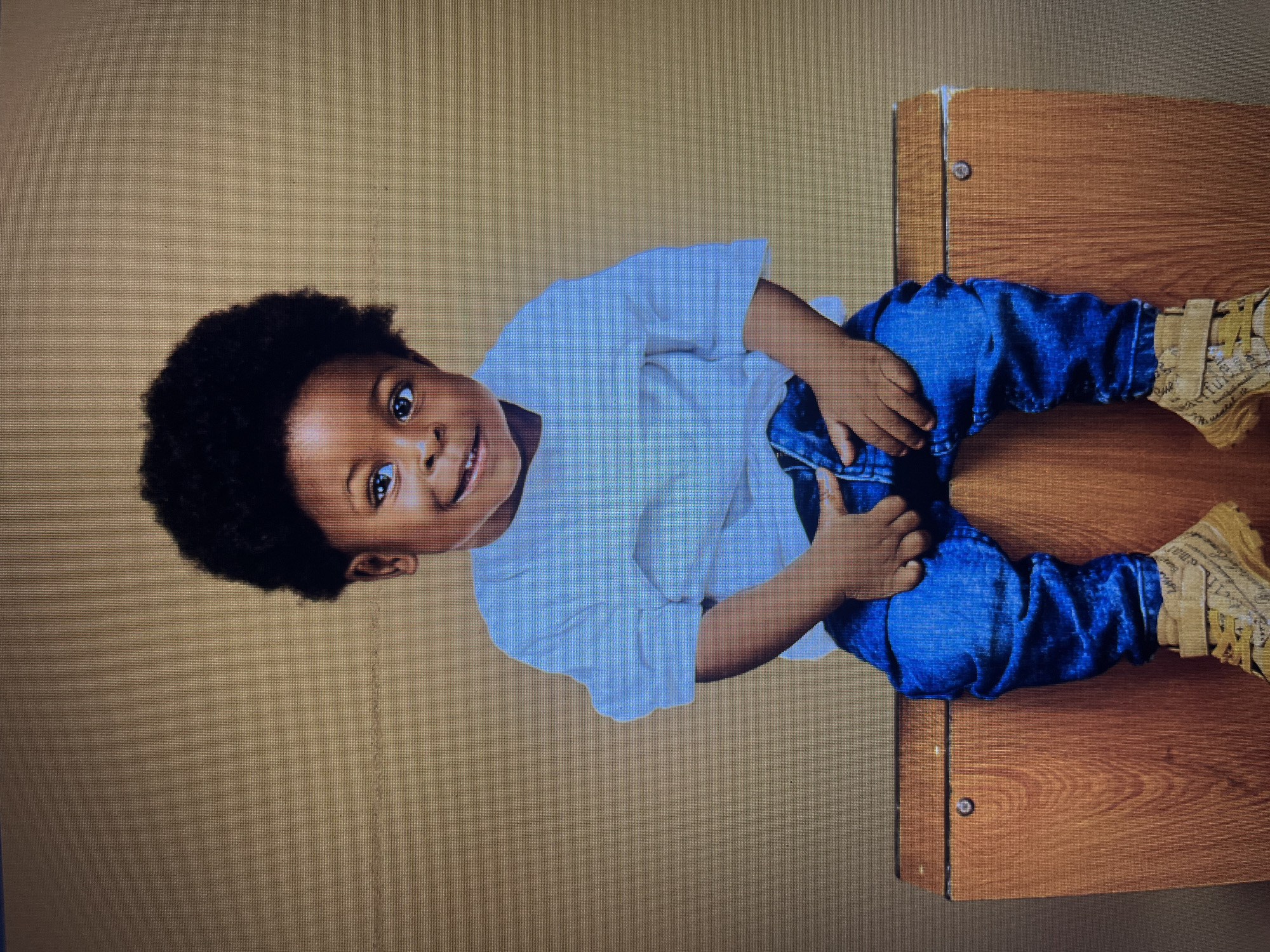 A young Black boy with a full afro is sitting on a wooden surface. He is wearing a white t-shirt and blue jeans. The boy is looking towards the camera and smiling. The background is a plain brown color. There is no information about the location of Gura Top, Nigeria in this image.