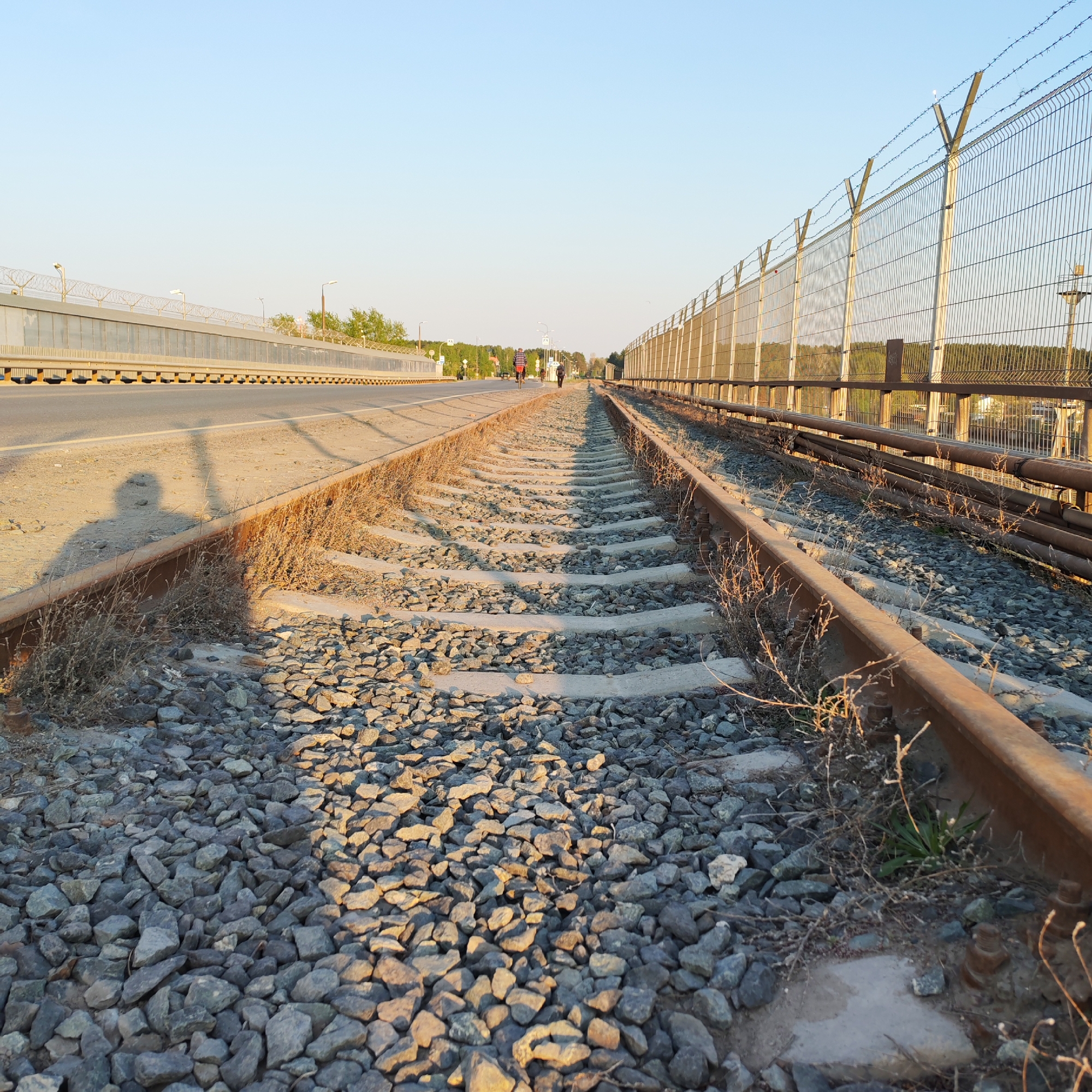 This image depicts a section of railway tracks in Chaikovsky, Russia. The tracks are bordered on one side by a highway and a bridge-like structure. On the other side, a tall chain-link fence topped with barbed wire runs parallel to the tracks. The ground around the tracks is covered in grey gravel and sporadic patches of dry vegetation. In the background, indistinct figures of people are visible on the highway. A long shadow is cast across the tracks on the left side, suggesting the presence of a person or object just outside the frame. The sky is clear and light blue, indicating daylight conditions.