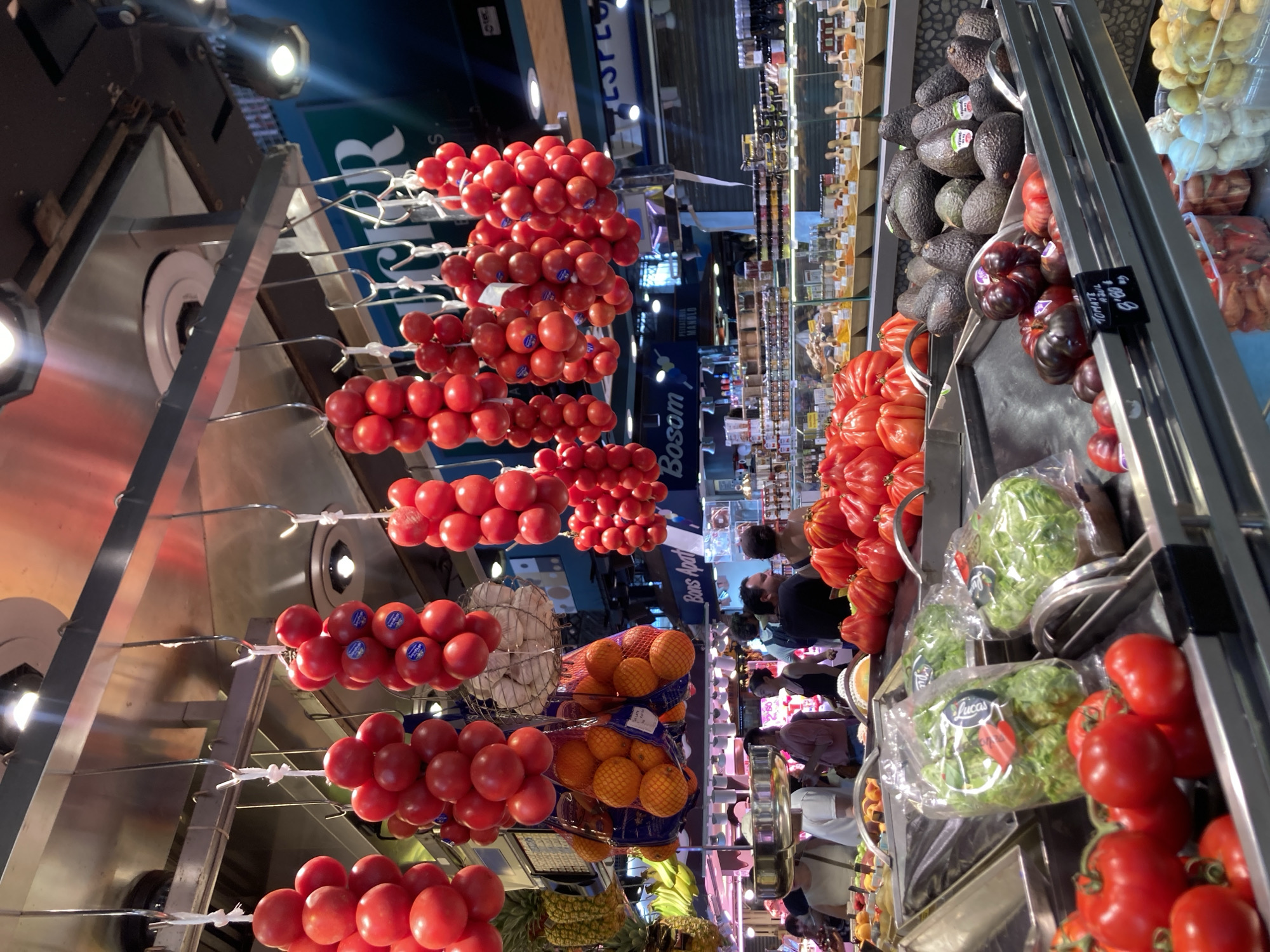 This media file captures a bustling scene inside a market, likely in Barcelona, Spain, given the location cue "Bosom Apart" and the general aesthetic of a European market. The primary subjects are a vibrant display of fresh produce, with a particular emphasis on tomatoes, which are presented in various forms: clustered on strings hanging from the ceiling, piled high in large piles, and some darker varieties with a price tag that reads "TOMATE 4,90 €". Avocados are also prominently featured, stacked in a large bin. In the background, several people are browsing and shopping, suggesting a lively atmosphere. The setting appears to be a well-lit indoor market with stalls displaying a variety of goods. The overall impression is one of abundance and freshness, typical of a food market. There are no clear indicators of the time of day or weather, but the indoor setting suggests it's regardless of external conditions. The mood conveyed is lively and active, with shoppers engaged in their purchases.