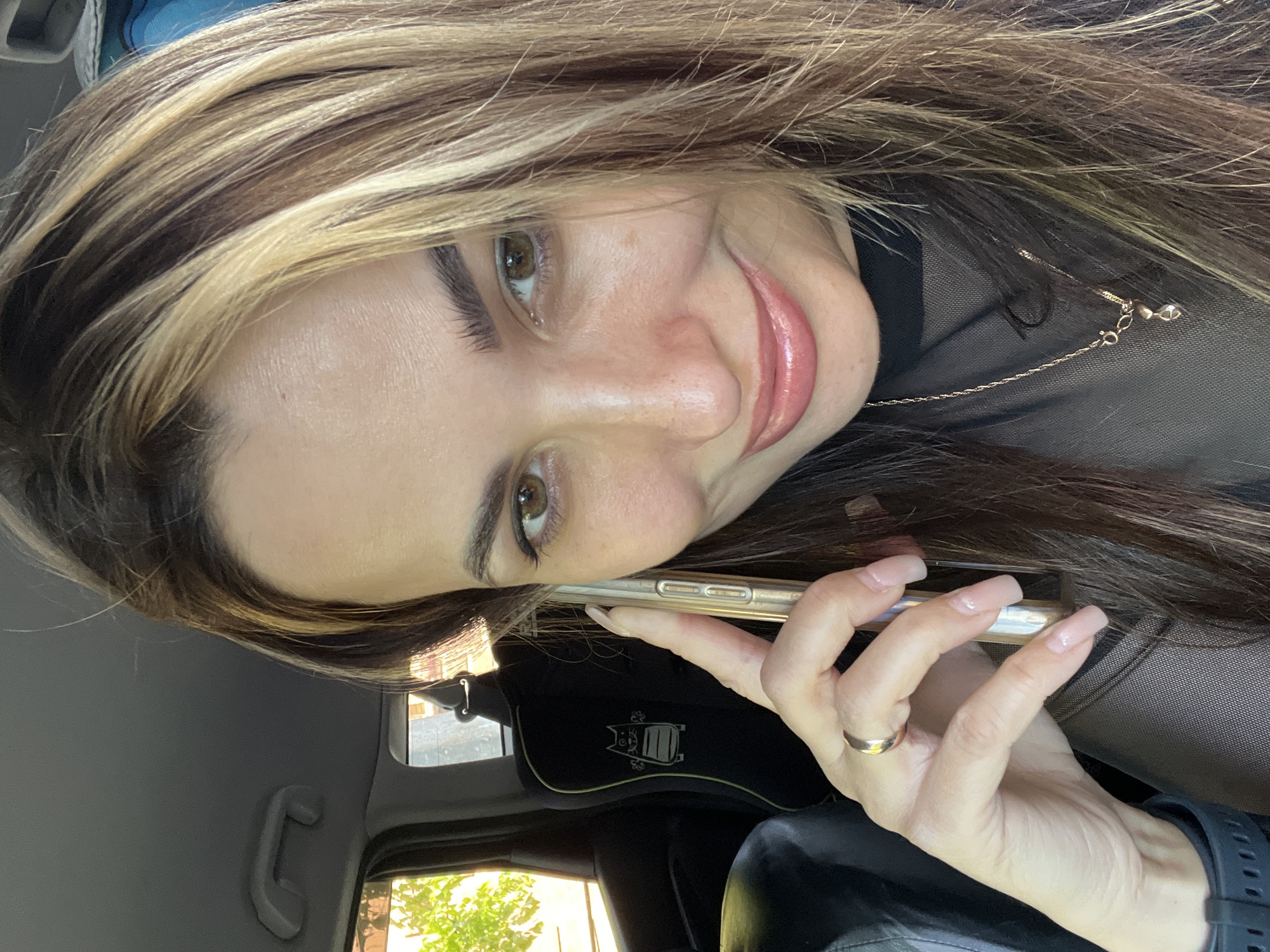 A woman is holding a smartphone to her ear, apparently engaged in a call. She is wearing a sheer, dark top with a gold necklace. She is inside a vehicle, evident from the car interior elements visible, such as a grab handle, window, and seat. The setting outside the car appears to be a residential area, as indicated by the glimpse of trees through the car window. The location is in the city of Unknown, Unknown.