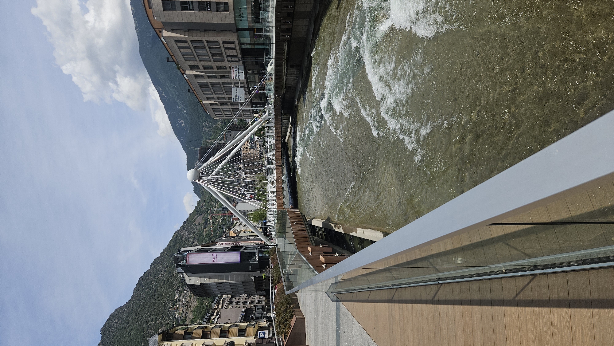 The image presents a detailed view of the Valira River flowing through Andorra la Vella, Andorra, on a bright, partly cloudy day. In the foreground on the left, a modern walkway composed of wooden planks is visible, bordered by a glass and grey metallic railing that angles towards the lower right corner.
The river, situated in the midground, exhibits clear, greenish-brown water with visible rocks and pebbles on its bed. It flows rapidly, generating numerous white rapids and turbulent sections. Spanning the river is a distinctive white pedestrian bridge, characterized by a central spherical mast from which multiple white cables radiate outwards to support its wooden deck. Large white letters spelling "ANDORRA LA VELLA" are prominently mounted on the bridge structure directly above the water.
Multi-story urban buildings line both banks of the river. On the left bank, structures include one with a vertical "Plan B" sign and a "P" parking sign. On the right bank, commercial buildings are visible, with a "MANGO OUTLET" sign partially discernible. The urban environment is set within a mountainous landscape, with steep, densely forested mountains rising in the background on both sides of the valley under a sky with scattered white clouds. No individuals are clearly discernible in the scene.