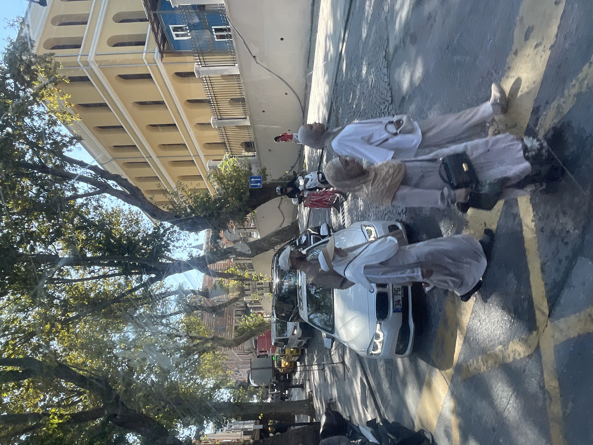 The image depicts a street scene in Istanbul, Turkey, with a view of a light yellow building with arched windows and a blue building with white-framed windows in the background. Trees with lush green foliage line the street. Several vehicles are present, including a white BMW sedan with a license plate reading "34 PG", a yellow taxi, and a white van. A man in a light-colored shirt and a white cap is standing next to the BMW. Three women, all wearing headscarves and long, flowing garments, are walking across the street, with two of them in the foreground. One of the women is carrying a black handbag. A person on a white scooter with a helmet is visible further down the street, alongside a red and white barrier. Another woman in a red top is walking on the sidewalk in the background. The street is paved with cobblestones and marked with yellow lines. The overall atmosphere suggests a sunny day in an urban environment.