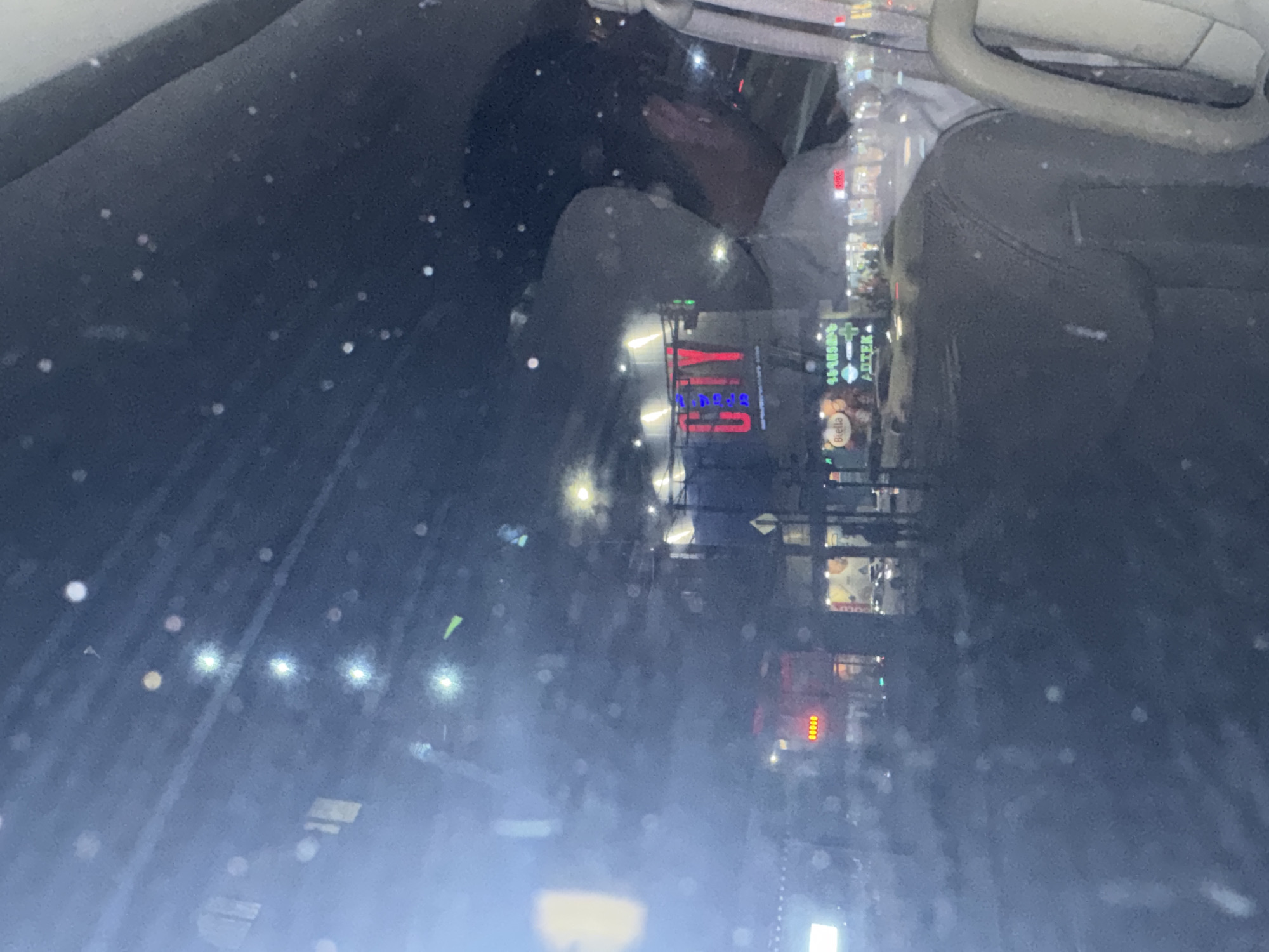 The image shows a scene inside a car at night. A person with dark hair is visible in the passenger seat, appearing to be sleeping or resting their head on their hand. The interior is dark, with raindrops or spots visible on the windshield. Through the windshield, reflections of city lights and signs are visible, including a sign with Cyrillic characters. Based on the signs and architectural styles, the location is Yerevan, Armenia. The street scene reflects commercial establishments and street lighting typical of an urban environment. The steering wheel and part of the dashboard are visible, indicating that the photo was taken from the driver's perspective.