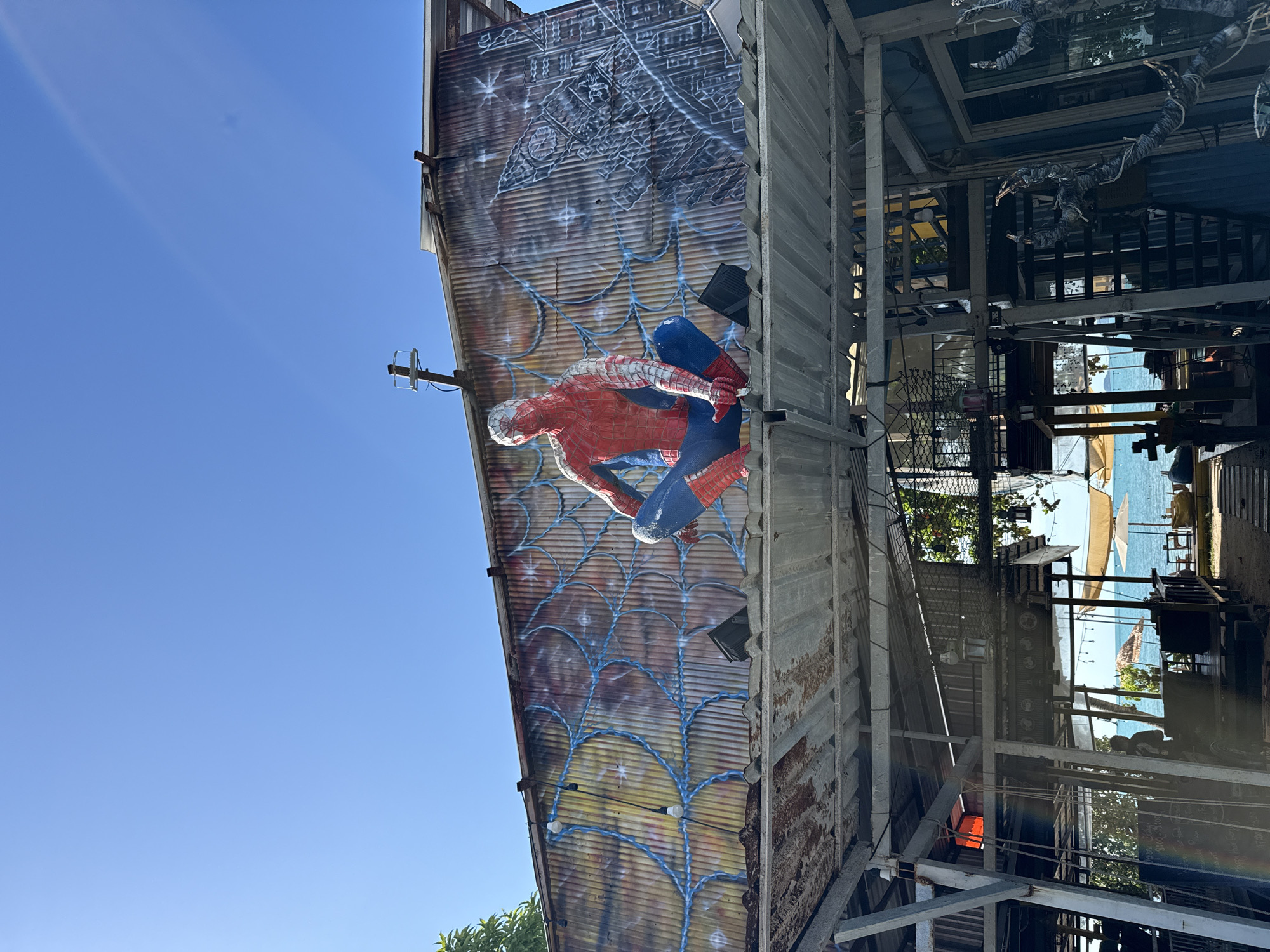 The image depicts a Spiderman mural on the corrugated metal wall of a building in Ko Samui District, Thailand, under a clear blue sky. The mural shows Spiderman in a crouching position, with a spiderweb design extending across the wall. To the right of the Spiderman mural, a cityscape is depicted in grayscale. Below the mural, the roof of a structure with exposed beams and corrugated metal sheeting is visible. In the foreground, the interior of an open-air establishment with metal framing, stairs, and various objects is shown. Further in the background, a beach scene with umbrellas and lounge chairs can be seen near the ocean. The overall scene suggests an outdoor location, possibly a restaurant or bar, with a playful artistic theme.