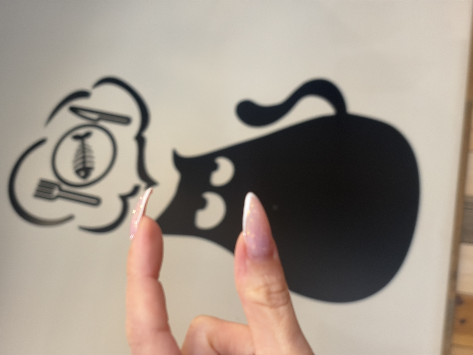 A close-up image shows a hand with manicured fingernails in the foreground. The background is a white surface with black graphic elements. One graphic depicts eating utensils (fork, knife) and a fish skeleton inside a cloud shape. Adjacent to this is a larger graphic depicting a black cat silhouette with a curled tail. The scene is in Saint Petersburg, Russia.