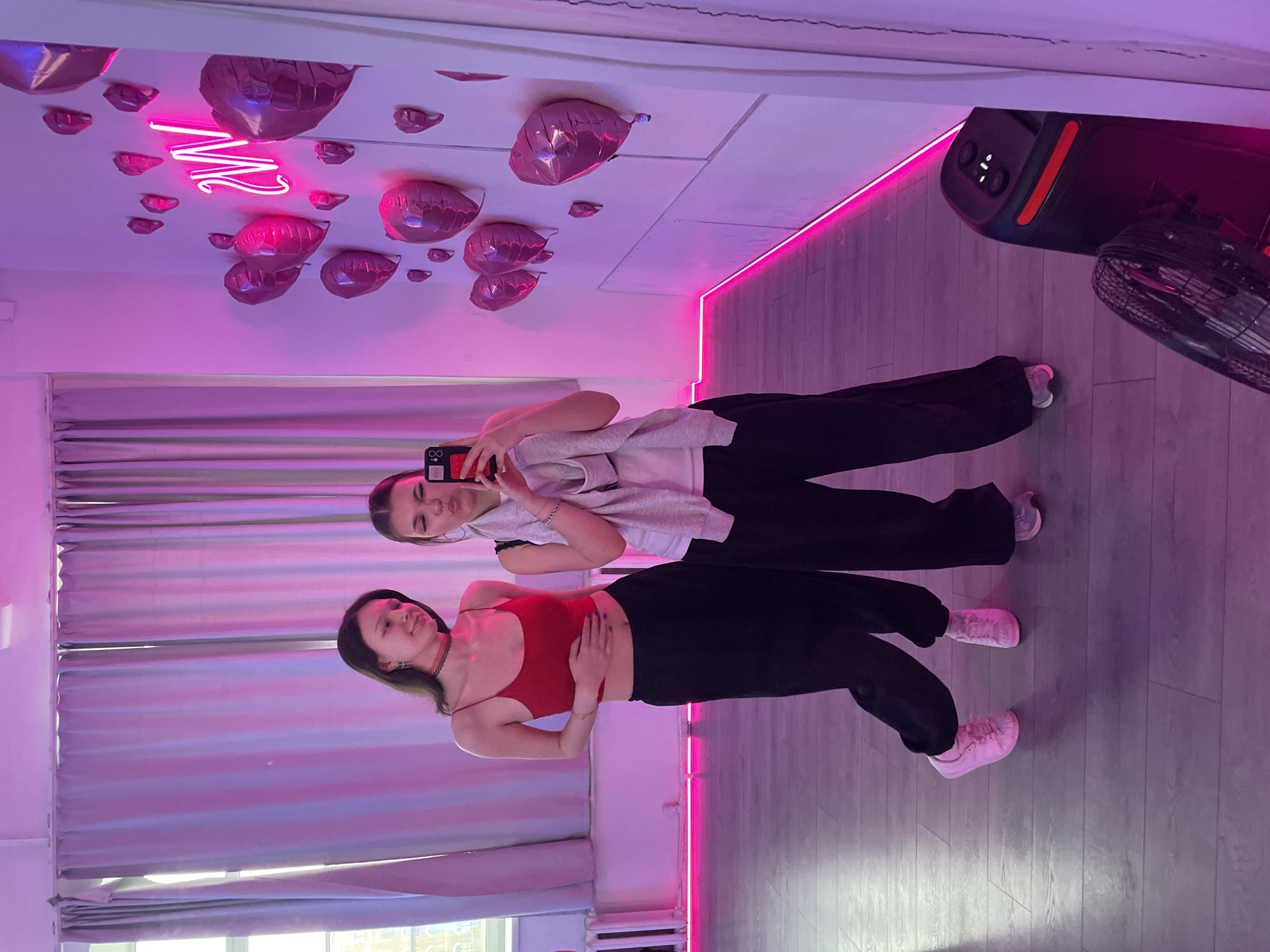 The image displays two young women posing in a room with vibrant pink and purple artificial lighting, primarily from LED strip lights running along the walls. The scene is dominated by a large mirror, reflecting both women and the room's decor.
In the reflection, the woman on the left wears a red crop top and black wide-leg pants, with her hands resting on her stomach. She has dark hair and a neutral, relaxed expression, looking towards the camera's reflection. Beside her, the woman on the right is taking a selfie using a smartphone, which is visible in her reflection. She is dressed in a light gray long-sleeve top layered over a dark shirt and black pants. Both women are wearing light-colored sneakers.
The room's ceiling, also visible in the mirror's reflection, is decorated with numerous pink heart-shaped balloons and features a glowing pink neon sign, stylized as a letter "M" or "W". Parts of a black sound system or speaker and a fan are also reflected in the upper right. The floor appears to be made of light-colored wooden planks. The smartphone case displays text that looks like "AXZ" and a camera icon. The overall ambiance suggests a casual photoshoot or a moment captured in a stylish, possibly event-oriented, indoor space. The artificial lighting indicates an indoor setting, likely in the evening or a space designed for specific ambiance, rather than daylight.