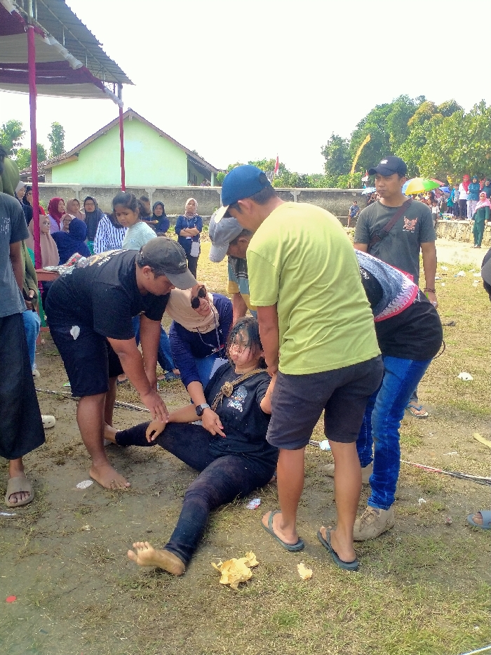 The image captures an outdoor scene in Celep, Indonesia, during daylight hours. A young adult female is positioned supine on a dirt and sparse grass surface, appearing unconscious or in distress. She is barefoot, wearing a black t-shirt and black leggings, with a braided cord around her neck. Her eyes are closed or partially closed.
Five individuals are clustered around her, providing attention or assistance. On her left, a male wearing a black baseball cap, black t-shirt, and black shorts is kneeling, with his left hand on her right forearm and his right hand near her shoulder. Behind him, a female wearing a beige headscarf, dark blue long-sleeved shirt, and blue jeans is also kneeling, with her right hand on the central female's left arm. On the central female's right side, a male in a blue baseball cap, light green t-shirt, and dark shorts stands observing. Another male, wearing a grey baseball cap, is partially visible leaning over from the right. A female in a black t-shirt with patterned sleeves and blue jeans stands to the far right, observing the situation.
In the mid-ground, a crowd of approximately two dozen individuals, including adults and children, observe the scene from a distance. Many female spectators are wearing headscarves. The background features a light green building with a reddish roof, a concrete wall, and dense green trees. To the far right, some individuals in the background crowd are standing under colorful umbrellas. A temporary white canopy structure with red and white support poles is visible on the left side of the frame. A crumpled, light-colored object, possibly paper or fabric, is present on the ground near the central female's feet.