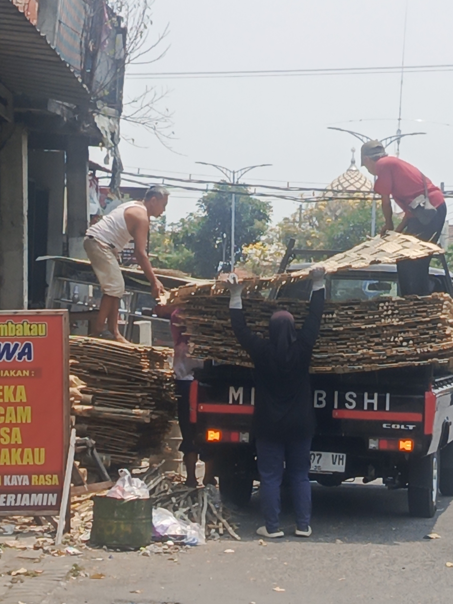 In Surabaya, Indonesia, under a bright sky, two men and a woman are engaged in the activity of loading a pickup truck with stacks of light brown, rectangular objects. The man on the left, wearing a white tank top and light-colored shorts, stands on a pile of these objects, reaching out to grasp another. The man on the right, clad in a red shirt and a cap, is positioned behind the truck, also handling the materials. The woman in the center, wearing a dark headscarf and blue trousers, is lifting one of the objects upwards. The objects appear to be wooden planks or similar building materials. A black Mitsubishi Colt pickup truck is central to the scene, with the word "MITSUBISHI" prominently displayed on its tailgate. The truck is laden with a significant quantity of these objects. The surrounding environment shows signs of an urban setting, with a building on the left exhibiting a corrugated metal roof and some hanging fabrics. In the foreground, on the left side, a red sign with yellow lettering is partially visible, displaying text that appears to be in Indonesian. A green barrel filled with what looks like trash is also present near the sign. The ground is paved, and scattered leaves suggest the presence of trees nearby. The overall scene depicts a labor-intensive task of material transport in an outdoor, commercial or industrial area of the city.