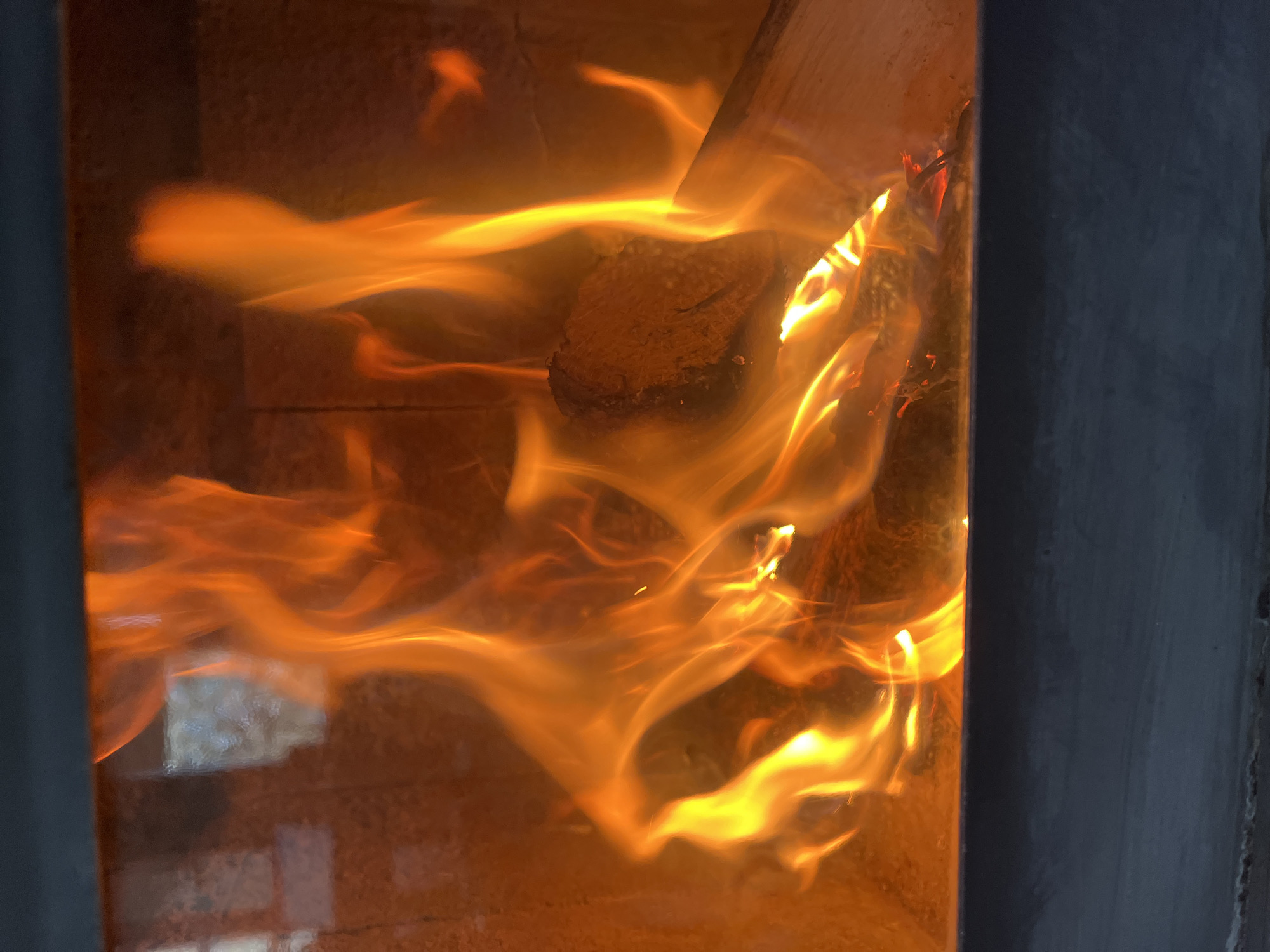 A close-up, slightly high-angle view focuses on a wood-burning stove in Pchelnik, Bulgaria. The stove is encased in dark grey metal trim. Inside, flames engulf pieces of wood and coal. The fire emanates an orange glow, creating a hazy background. The edges of brick can be seen behind the flames.
