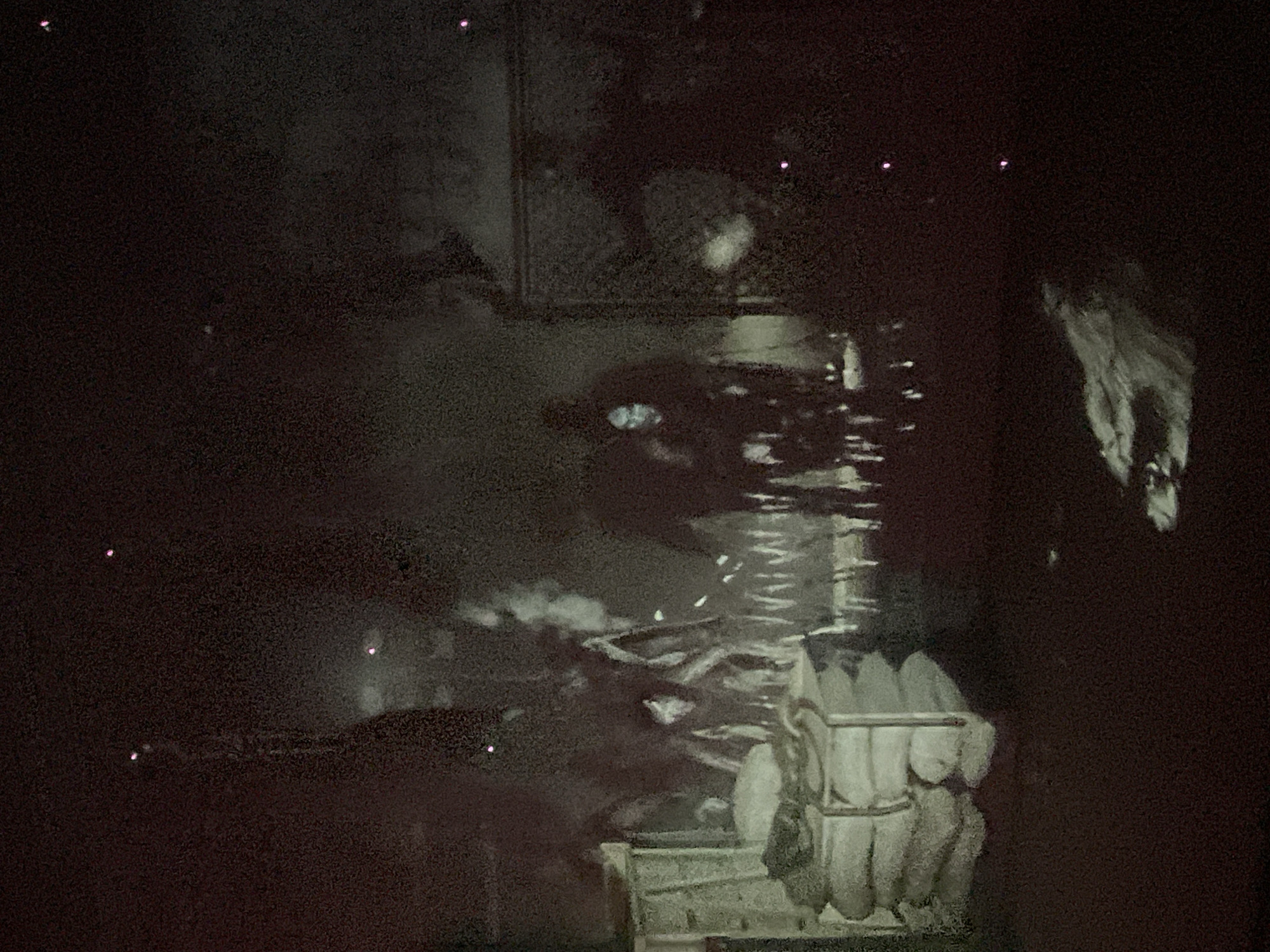 The image depicts a dimly lit, possibly indoor scene with several figures and objects. In the foreground left, there is a stack of what appears to be sacks or bundles, covered with a lighter colored material. To the right of these sacks, there is a fire or heat source producing a significant amount of smoke or mist. Several figures are visible within this smoky area; one in the center appears to be standing and looking forward, with a pale face. Another figure is to the left, partially obscured. The background is dark, with some indistinct structures or walls. Scattered throughout the scene are small, bright points of light, resembling distant lights or embers. In the lower right portion of the image, there is a light-colored object that appears to be a piece of debris or wood. The overall impression is one of a mysterious, possibly abandoned or dimly illuminated space.
There is no identifiable information within the image or the provided crops that suggests a specific location in the city of Denpasar, Indonesia.