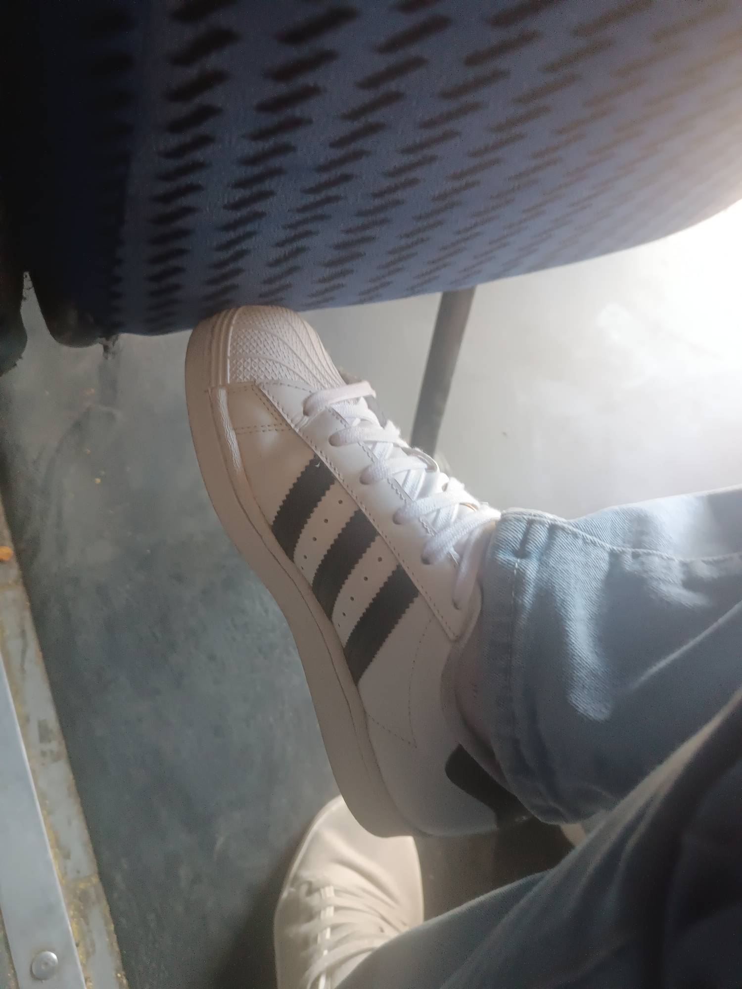 In Porlamar, Venezuela, a person wearing light blue denim jeans and white Adidas sneakers with black stripes is seated. The left foot is prominently featured, resting near a dark blue, textured seat. The surrounding environment appears to be an indoor space with a light-colored floor. The lighting suggests a bright day, possibly with sunlight entering from the right side of the frame, creating a hazy effect.