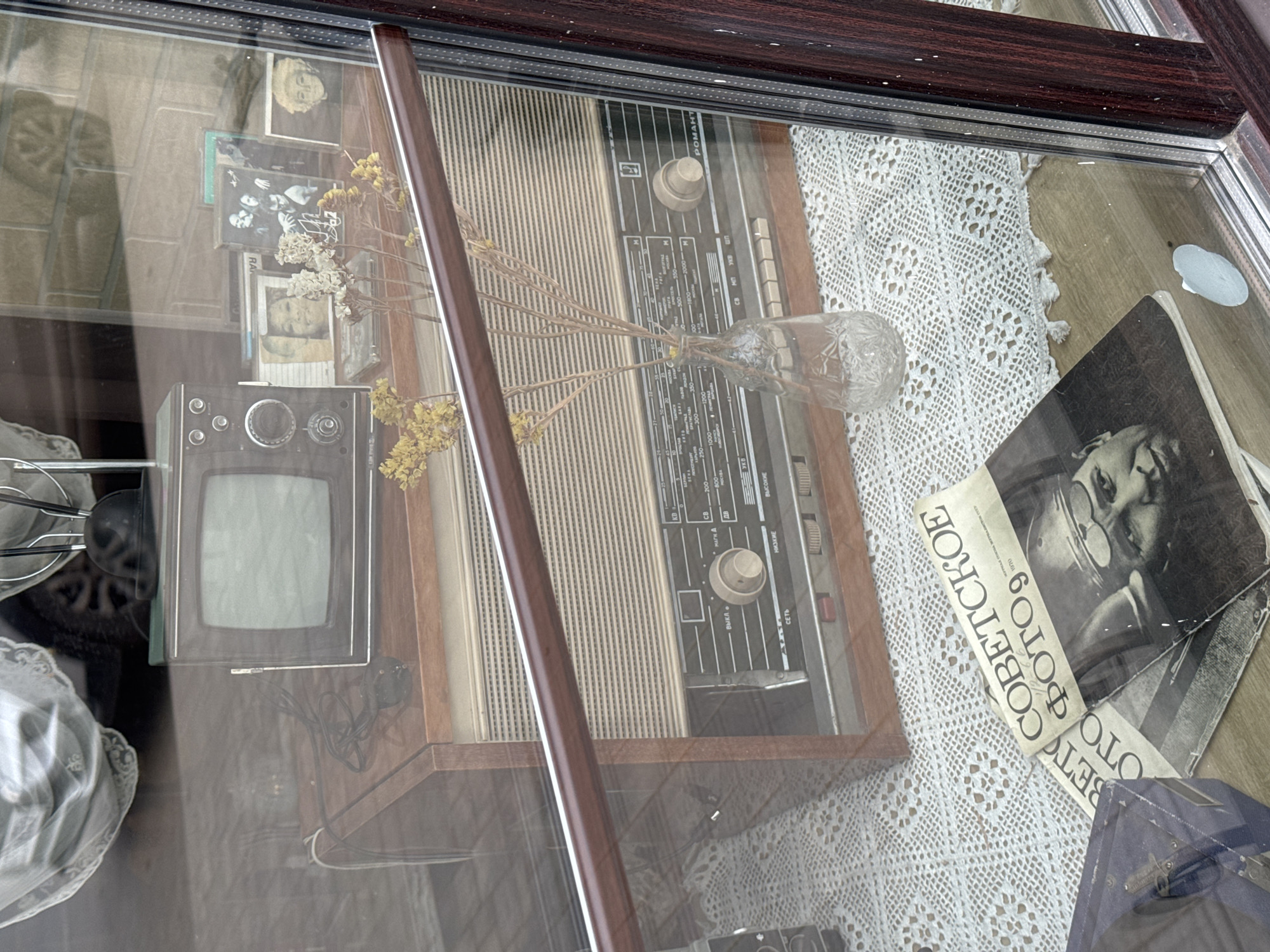 The image shows a display of vintage objects behind glass, likely in a store or exhibition. There's a small, old-fashioned television set, a radio with a frequency dial, a clear vase containing dried flower stems, some photographs, and a piece of lace fabric. The text on one of the photographs appears to be Russian: "СОВЕТСКОЕ ФОТО 9" (Soviet Photo 9). The radio has "РОМАНТ" ("ROMANT") written on the front. The image is taken from an elevated angle, looking down into the display. There are reflections visible on the glass. The exact location in the city of Kladovitsy, Russia cannot be precisely determined from the image alone.