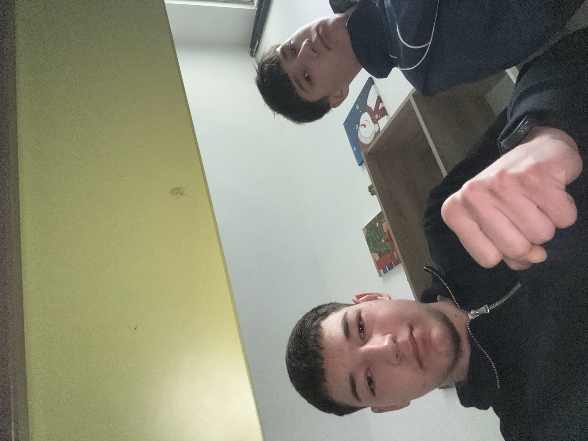 Two young men are visible in the image. One is wearing a black hooded jacket, and the other is wearing a dark blue jacket with white trim. A partially visible shelf unit is against a white wall, adorned with two small pictures. There is a yellowish wall visible on the left side of the frame. The setting appears to be indoors, potentially in a residential area, although the exact location in Zlatograd, Bulgaria cannot be determined from the given image.