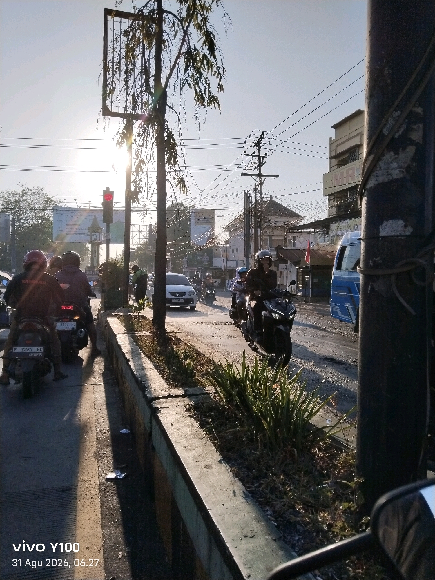 The image captures a street scene in Situbondo, Indonesia, during daylight. The sun is low in the sky, casting long shadows. In the foreground, a motorcyclist wearing a red helmet is positioned on the left, with another motorcyclist behind them. To the right, a person is riding a motorcycle with a blue body. Further down the street, a white car is visible, and several other motorcycles are on the road. The street is lined with a concrete barrier and vegetation. In the background, buildings and power lines are present. A traffic light is illuminated red. A large billboard with text is also visible in the distance. The timestamp in the lower-left corner indicates "vivo Y100 31 Agu 2026, 06.27".