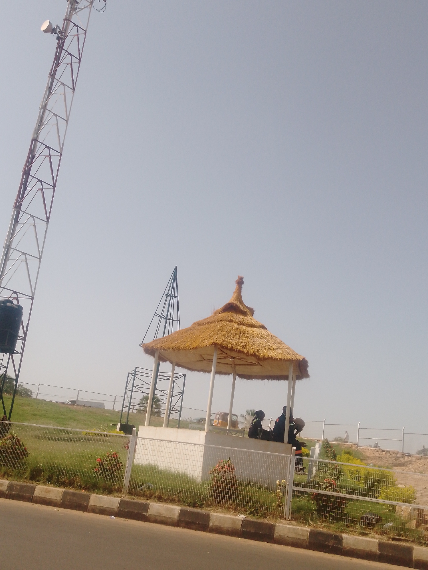 The image shows a scene in Sabon Tasha, Nigeria. A tall, red and white communication tower is prominent on the left. A small, thatched-roof gazebo stands in the center, with three people visible sitting on the wall under it. A white fence surrounds a grassy area with some bushes and flowers. Another metal structure with a peaked top is behind the gazebo. A road is visible in the foreground. The sky is light blue.