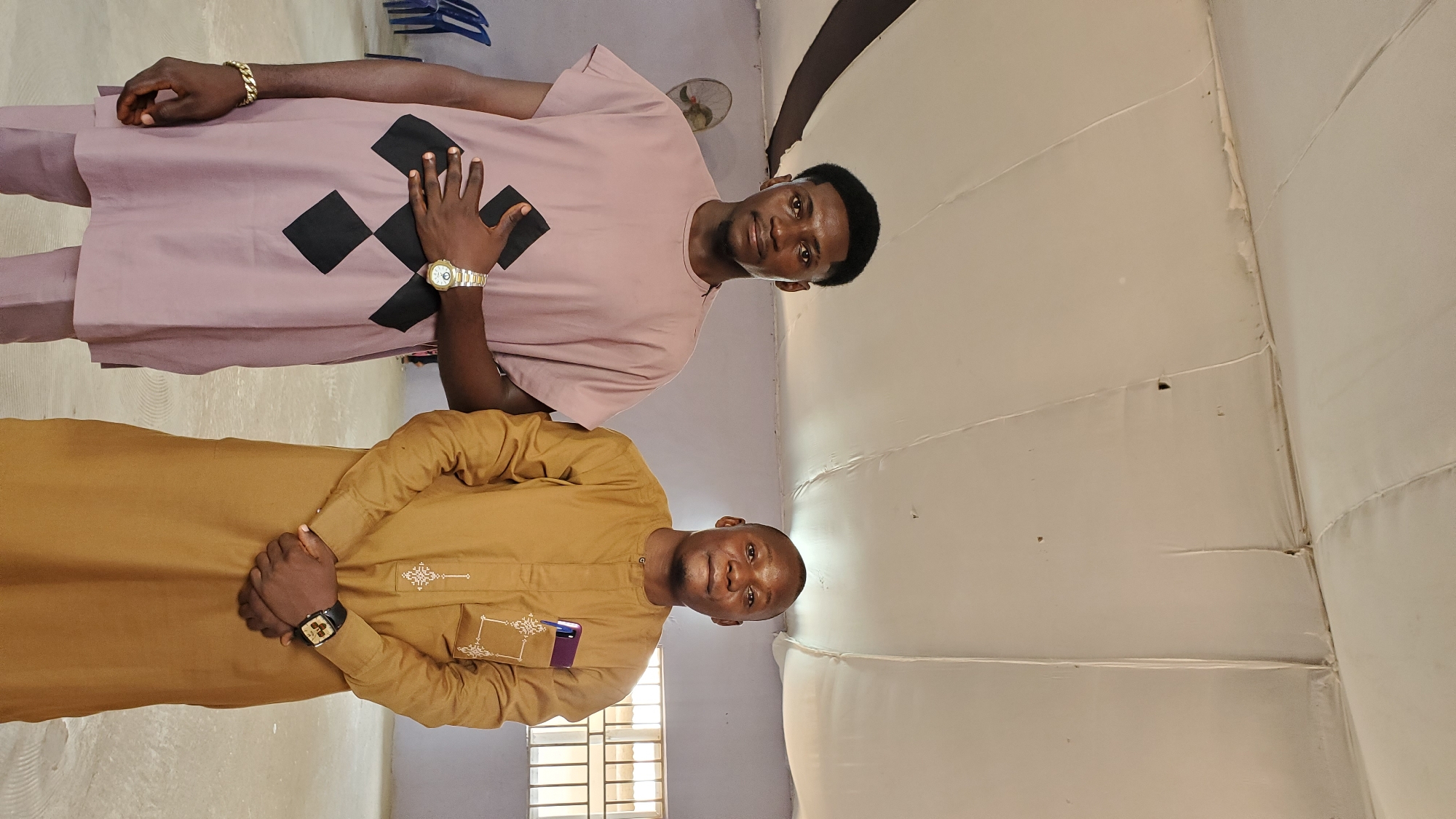 Two adult males stand side-by-side indoors, posing for a photograph. The male on the left wears a light pink traditional outfit with black diamond patterns on the chest and gold jewelry on his wrist. He has dark hair and is looking directly at the camera. The male on the right wears a tan traditional outfit with embroidered details on the chest pocket and a watch on his left wrist. He also has dark hair and is looking at the camera with a slight smile. They are in a room with a white tent-like ceiling and pale walls. In the background, a fan is mounted on the wall, and there are chairs visible. A window with metal bars is also present in the background. The scene appears to be a public space or an event venue in Jalingo, Nigeria.