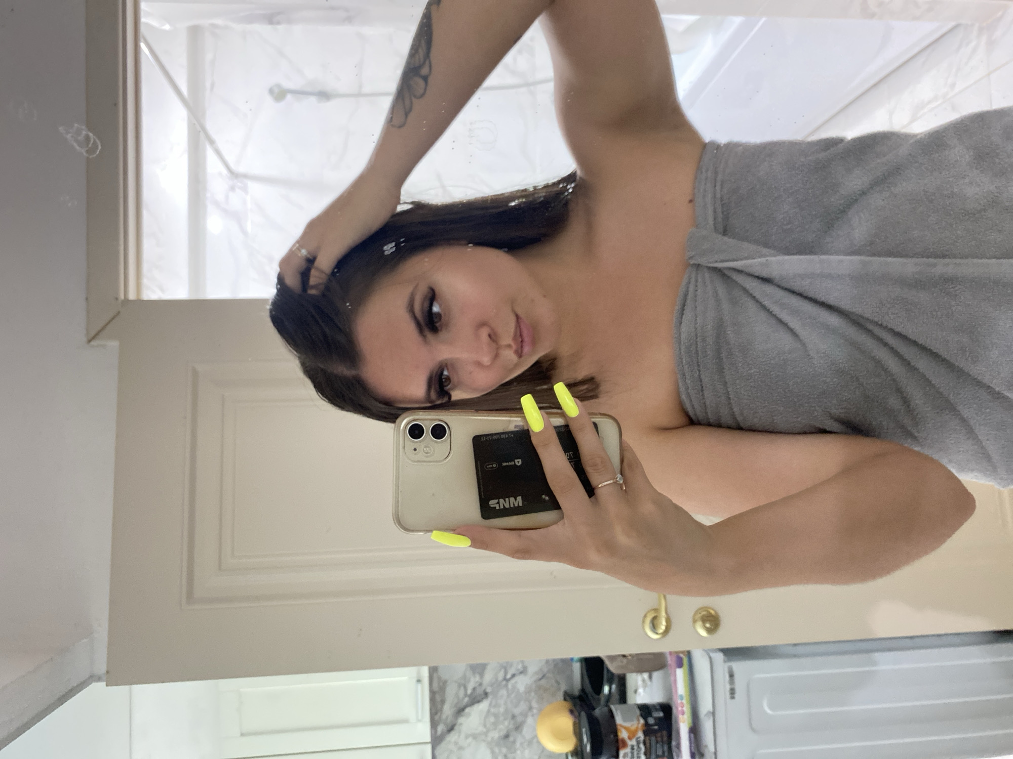 The image is a vertical, slightly blurry selfie taken in a bathroom mirror. A young woman, with long dark hair and visible makeup, is wrapped in a gray towel. She is holding her phone in her right hand, taking the picture, and her left hand is raised to her head. Her nails are painted a bright neon yellow. The background shows a tiled shower curtain and a white door. Visible text on the phone case reads "SNM" and some numbers that appear to be a card number. The overall impression is casual and personal, possibly taken after a shower.