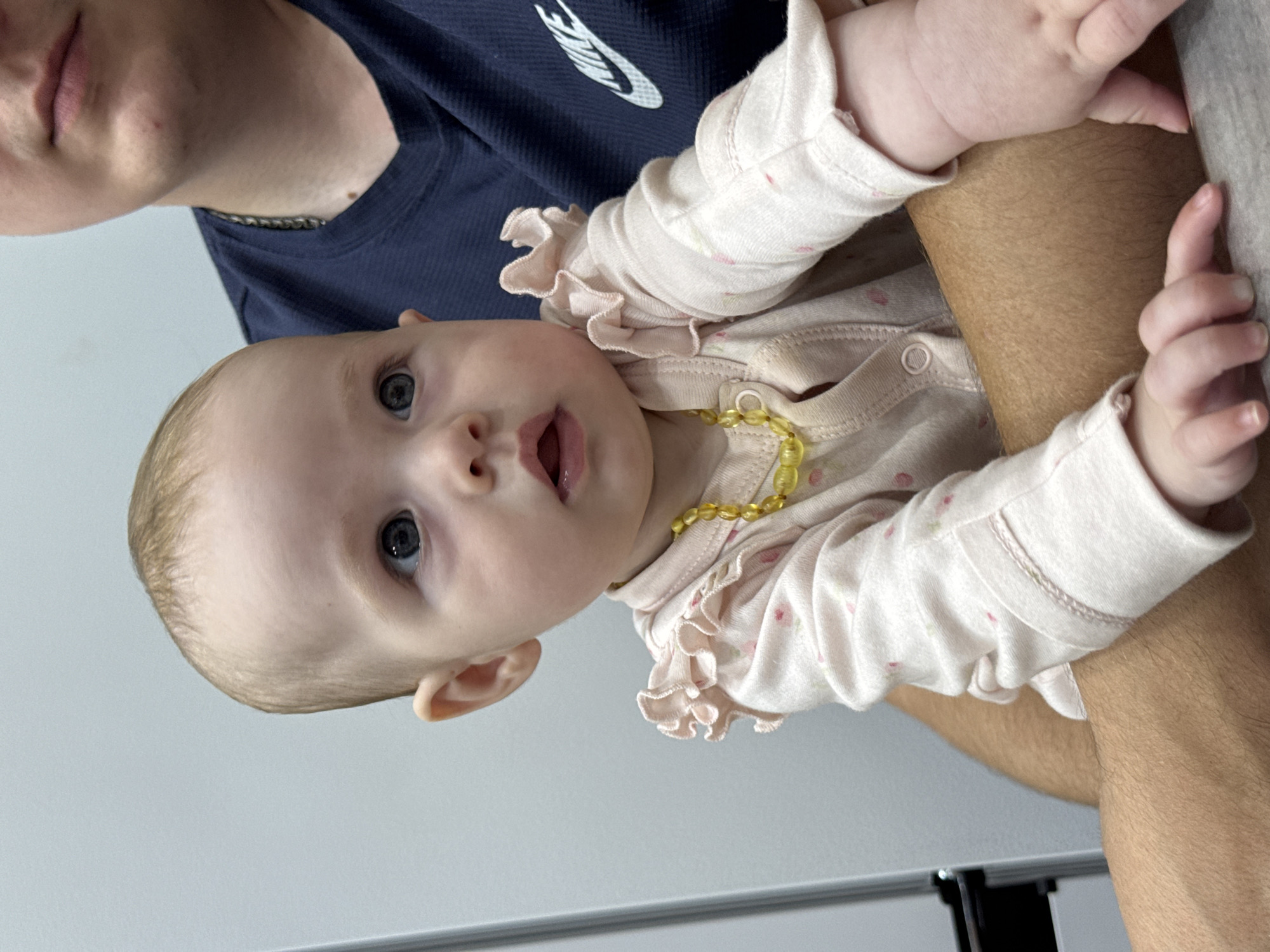 A close-up, slightly angled shot shows a baby held by an unseen person. The baby, with fair skin and light brown hair, is wearing a pink outfit with small pink dots and ruffled sleeves. A yellow necklace is around the baby's neck. The baby's eyes are open and looking to the left. The background is a plain, light-colored wall. The scene appears to be indoors. There are no specific geographical indicators in the image that would suggest the location is Noviy, Russia.