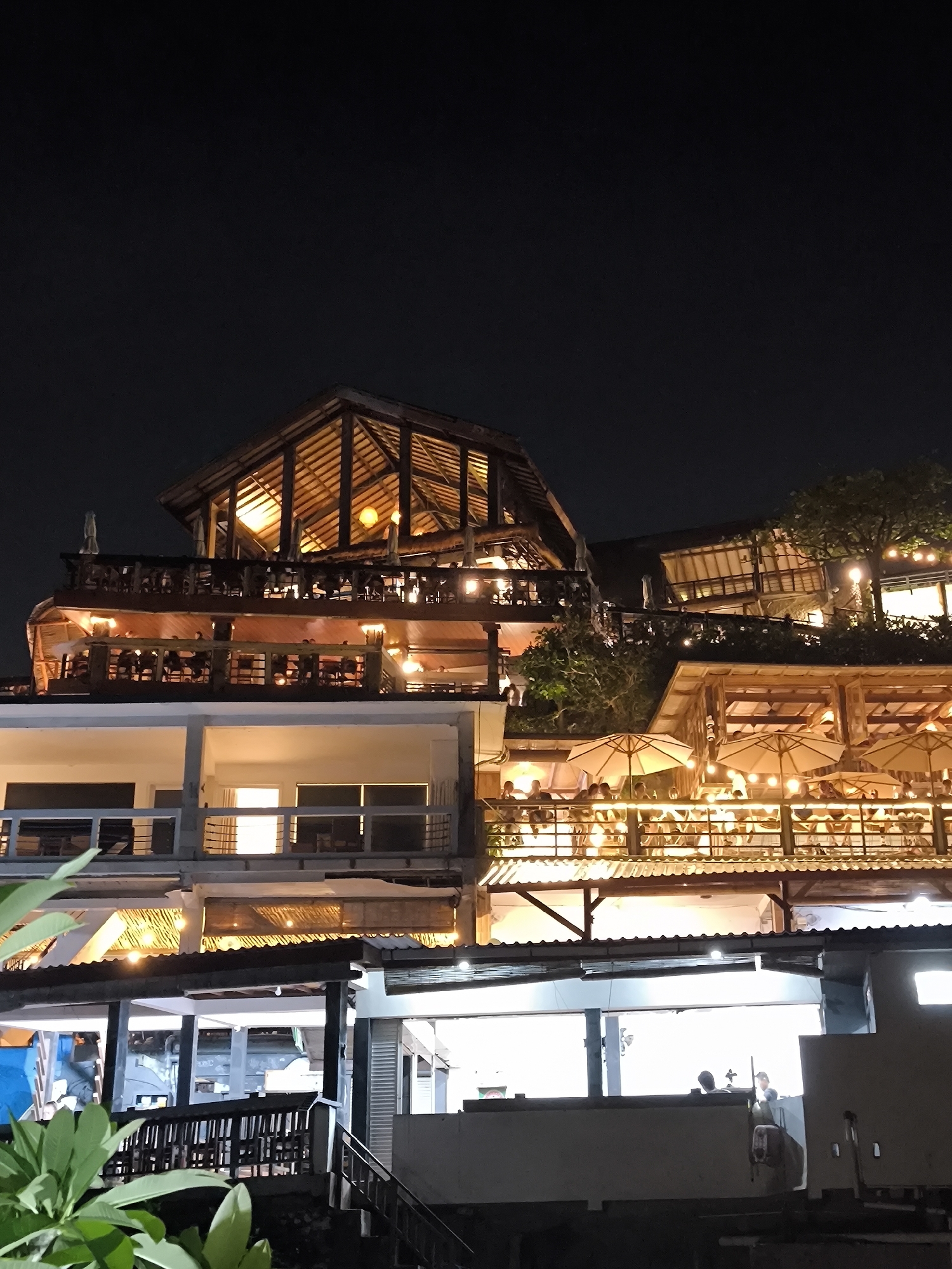 At night, a multi-level restaurant complex is brightly lit in Uluwatu, Indonesia. The architecture incorporates wood and open-air designs. People are visible on different levels, some seated at tables under umbrellas. A tree is interspersed between the structures. The sky above is dark. A staircase leads down towards the bottom levels, obscured by greenery and shadows. The complex is built into a hillside, with different levels cascading downwards.