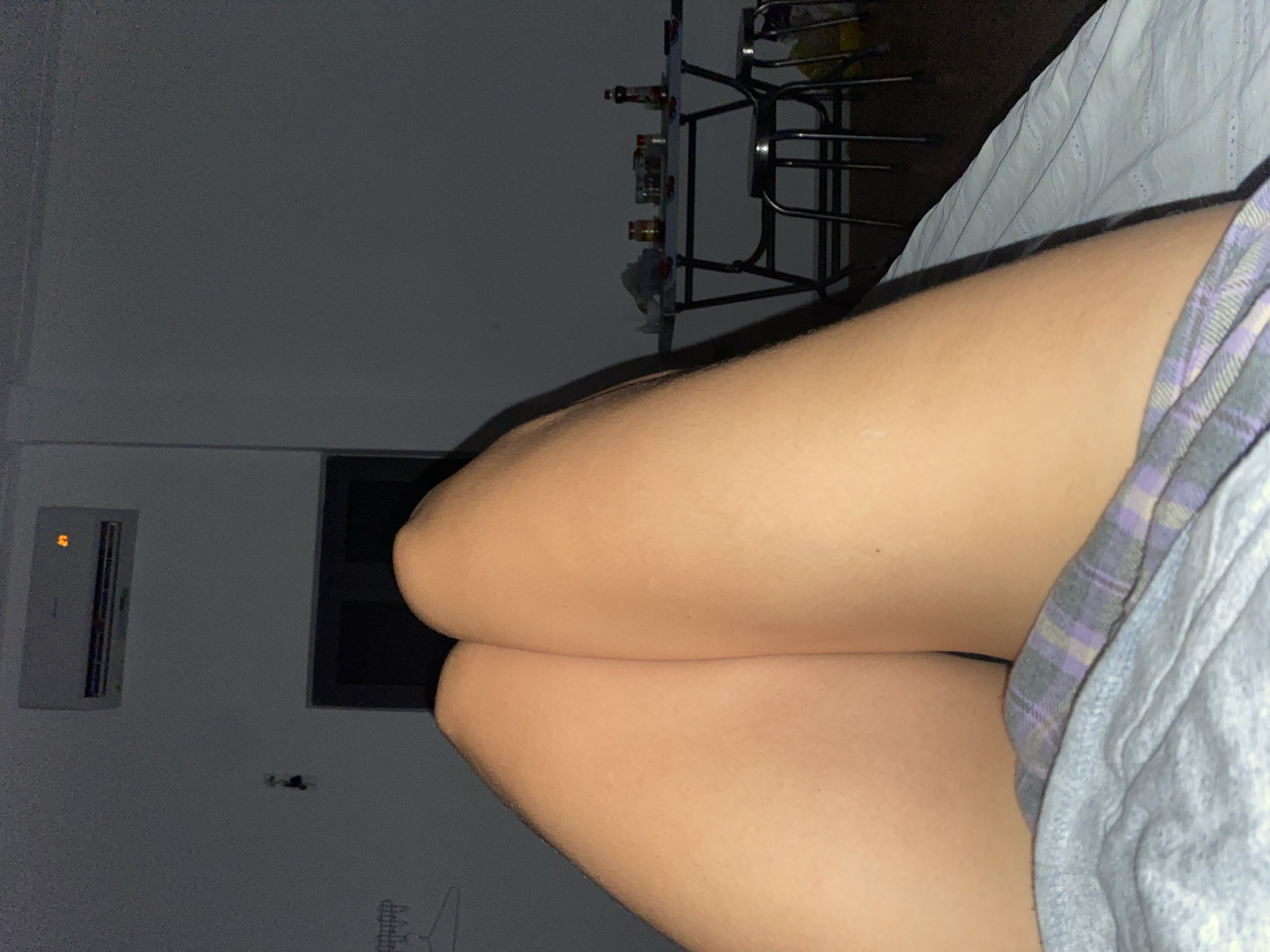 The media file shows a close-up, side-view of a person's legs, specifically the thighs and knees, indoors. The person is wearing a plaid pajama-like garment on their right leg, which appears to be purple and grey. The skin on the legs is light-skinned. The lighting is dim, suggesting it is nighttime or a dimly lit room. In the background, there is a glimpse of a small table with several bottles on it, possibly a bar or a party setup. To the left of the frame, a white air conditioning unit is mounted on the wall, displaying a faint orange light. Further to the left, a dark rectangular shape, likely a window or a doorway, is visible. The overall scene suggests a relaxed, indoor setting at night. There is no visible text in the image.