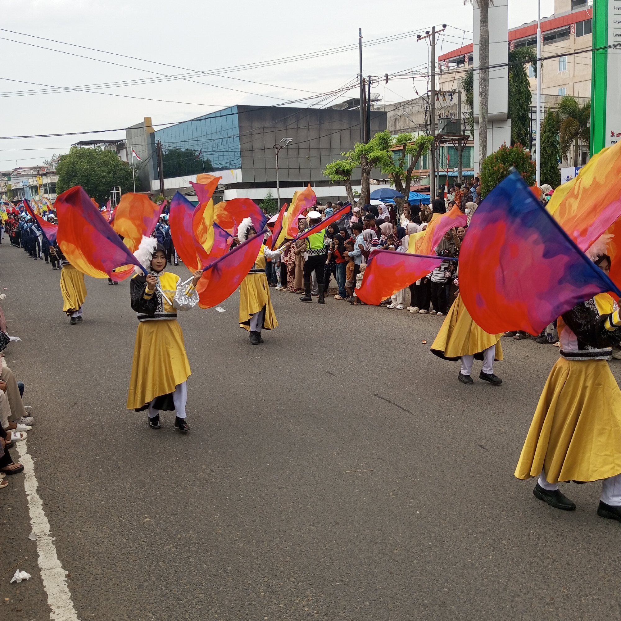 The image depicts a street parade in Lhokseumawe, Indonesia. A group of young women in matching yellow and black uniforms are performing with large, colorful flags that transition from red to orange to yellow and blue. They are marching on a paved road, with spectators lined up on the sidewalk. Buildings, including a modern structure with a blue glass facade, and utility poles are visible in the background. The sky is overcast. The overall scene suggests a festive or cultural event taking place in the city.