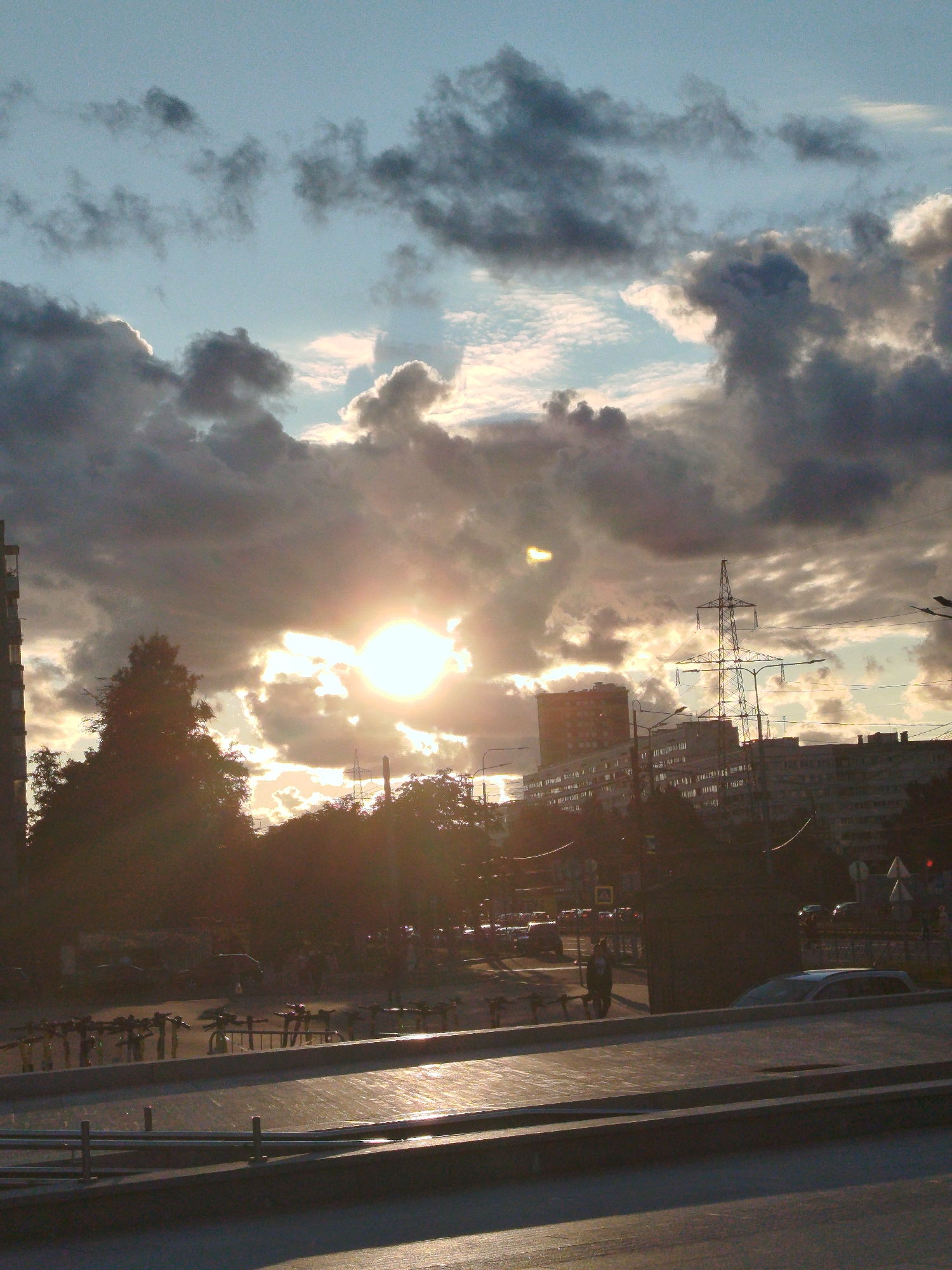 The image depicts a city scene in Saint Petersburg, Russia, during a sunset. The sun, a bright orb, is partially obscured by dark, dramatic clouds, casting a warm, golden light over the scene. The sky is a mix of blue and gray, with wispy white clouds scattered amongst the darker ones.
In the foreground, there is a concrete structure with a metallic railing. Beyond this, a street level area is visible with a row of parked bicycles. Further back, a line of trees, silhouetted against the bright sky, partially hides buildings. One tall apartment building is prominent on the left, with many windows.
In the midground, a large electrical pylon stands tall, with its crisscrossing wires visible. More multi-story buildings, typical of urban Russian architecture, are present. There are streetlights and traffic lights visible, indicating a populated area. A few cars are on the road, and some people are walking. The overall impression is of a busy urban environment experiencing the end of a day with a striking sky.